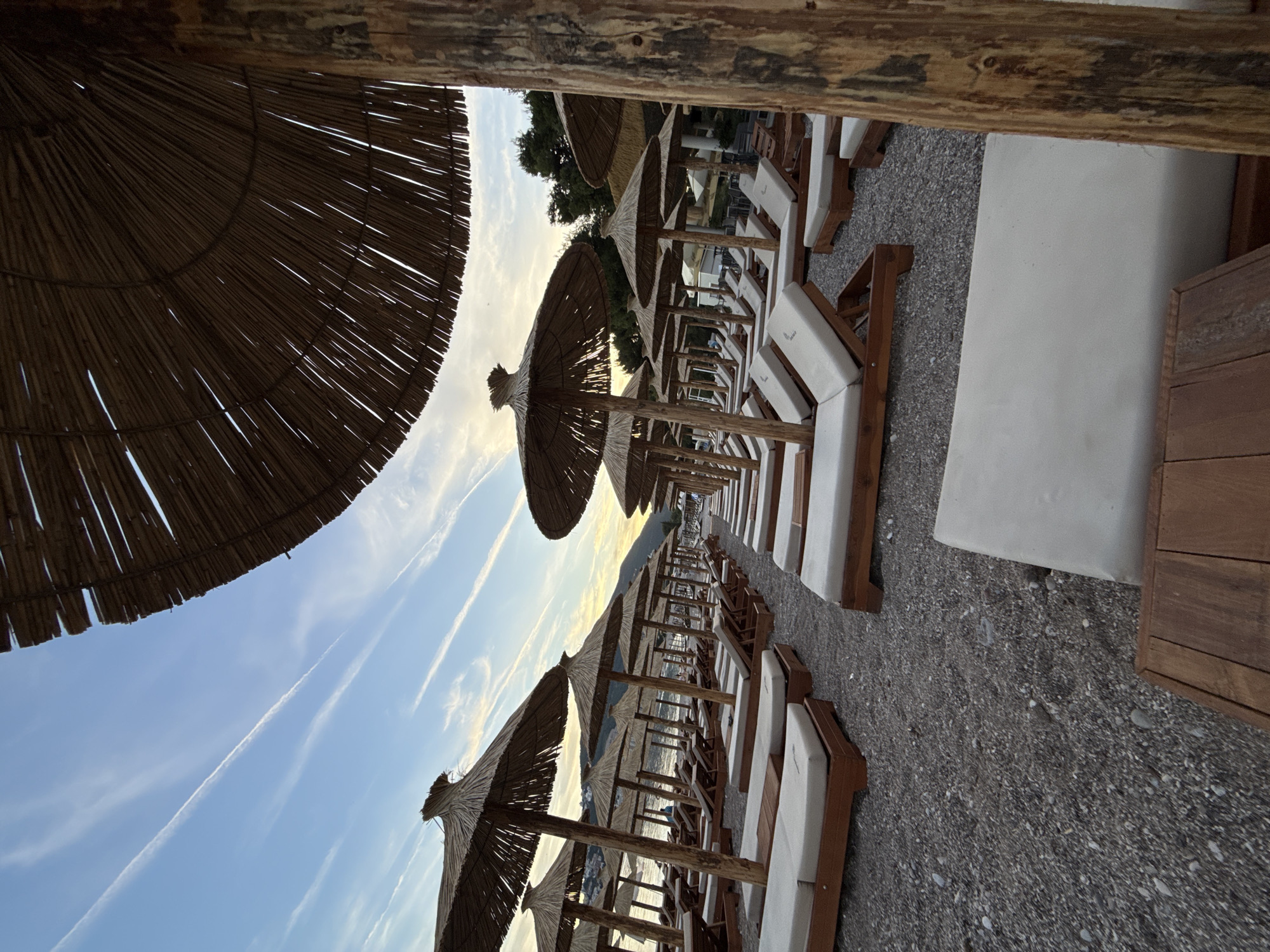 The image depicts a beach resort in Budva, Montenegro, during twilight. The foreground is dominated by rows of empty, white-cushioned sun loungers and thatched umbrellas, suggesting the end of the day or an off-season period. The beach itself is covered in dark grey pebbles. In the background, the sea is visible, with a faint outline of hills or mountains on the horizon. The sky above is a pale blue with streaks of white clouds, likely contrails from aircraft, indicating civil aviation activity. The overall atmosphere is serene and tranquil.