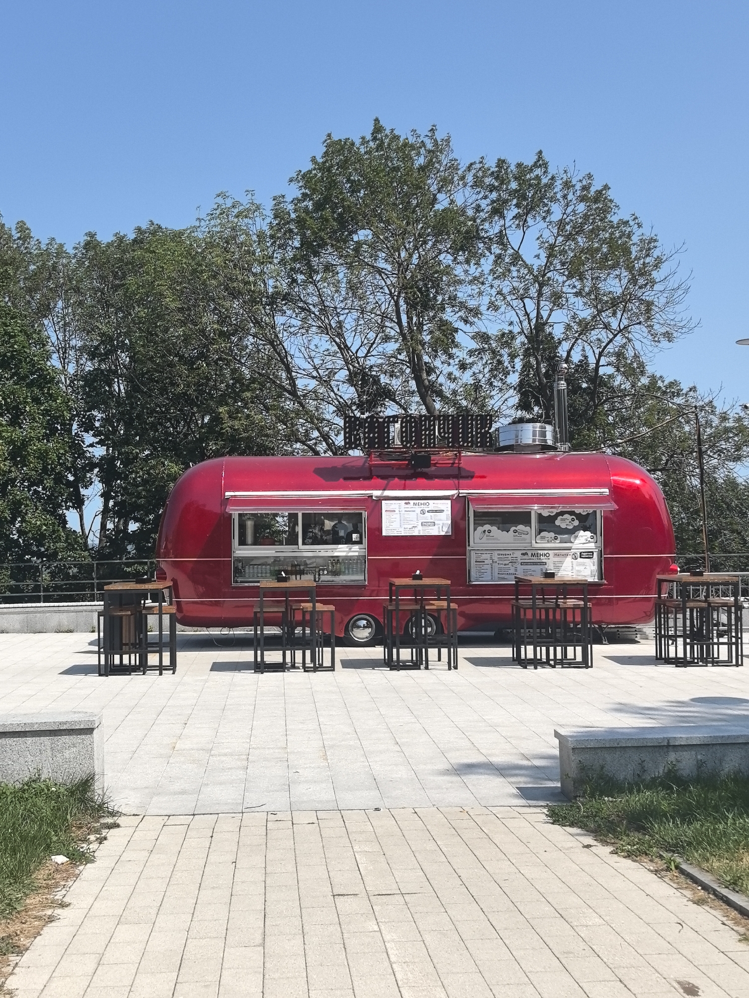 The image shows a bright red, vintage-style food truck with tables and chairs set up outside on a paved area. The food truck is shiny and appears to be a popular spot, with a menu visible in one of its windows. Several trees with lush green leaves are visible in the background, along with a clear blue sky. The overall setting suggests a pleasant, outdoor dining experience, possibly in a park or recreational area. The food truck has the name "MEHIO" prominently displayed on its menu board. The food truck is situated on a modern, tiled plaza, with some greenery and stone benches visible. The scene is bright and sunny, indicating daytime. This location is most likely the Nizhny Novgorod Kremlin embankment, known for its food vendors and recreational spaces.