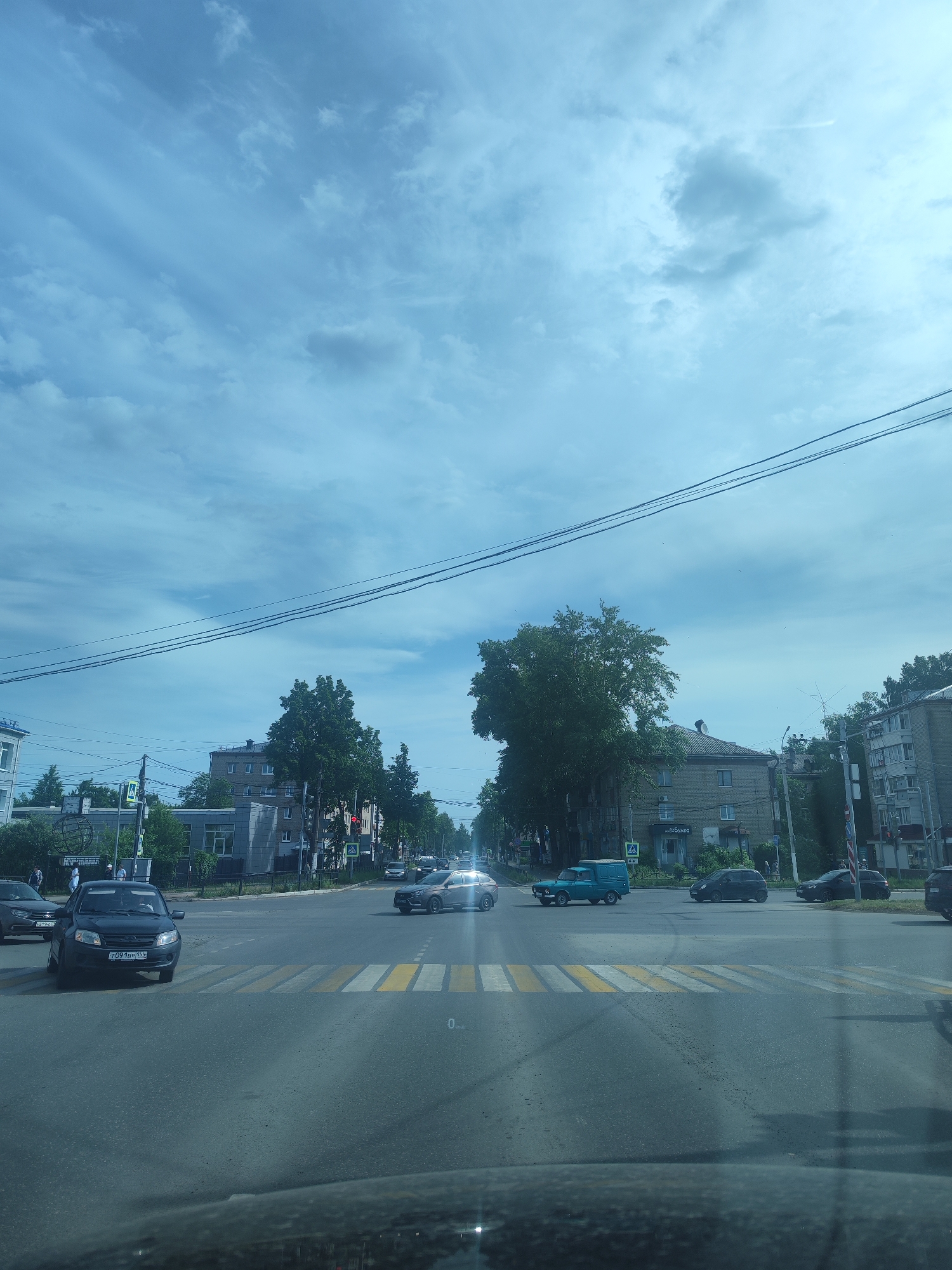 A street scene in Chaikovsky, Russia is depicted during daylight hours under a cloudy sky. An intersection is visible with several vehicles present, including a dark-colored compact car with the license plate "T091BP 159" stopped at a crosswalk, a silver sedan moving through the intersection, and a light blue van in the distance. Buildings line the streets, featuring a mix of residential and commercial structures, including a business with the sign "БУЛКО". Overhead, power lines stretch across the sky. The viewpoint suggests the photo was taken from inside another vehicle, positioned on the road leading to the intersection. There are zebra markings on the pedestrian crossing in the road.