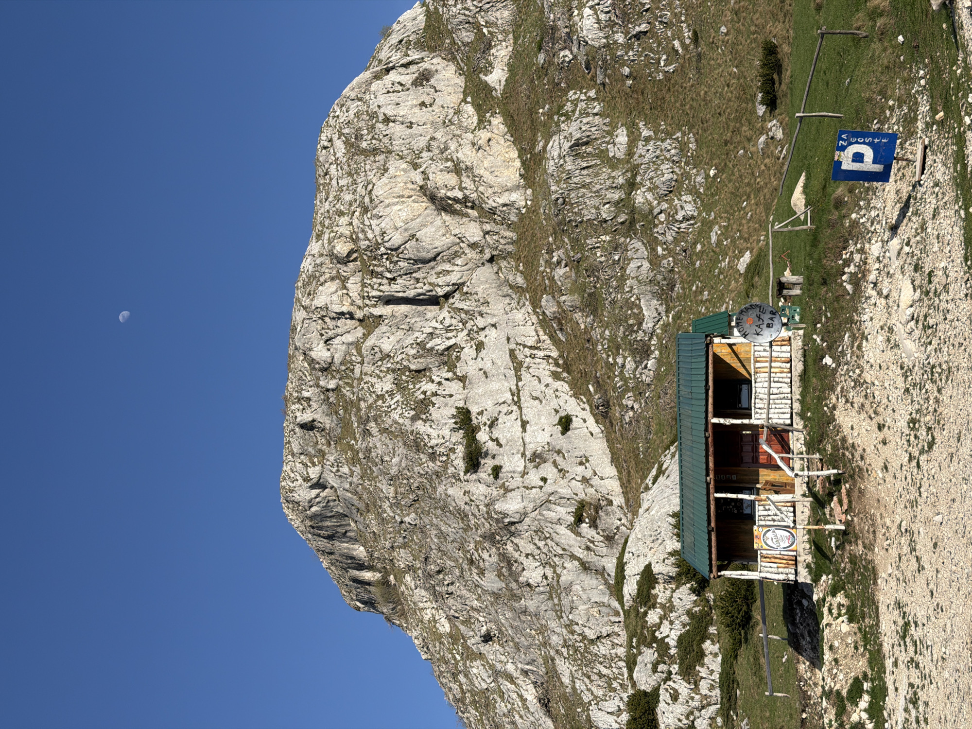 The image depicts a mountain scene in Boričje, Montenegro, featuring a rocky mountain face under a clear blue sky with a visible crescent moon. A wooden structure, likely a mountain hut or cafe, sits on the mountainside, adorned with signs, including a "parking" sign in Cyrillic. The surrounding terrain is a mix of rock and sparse vegetation.