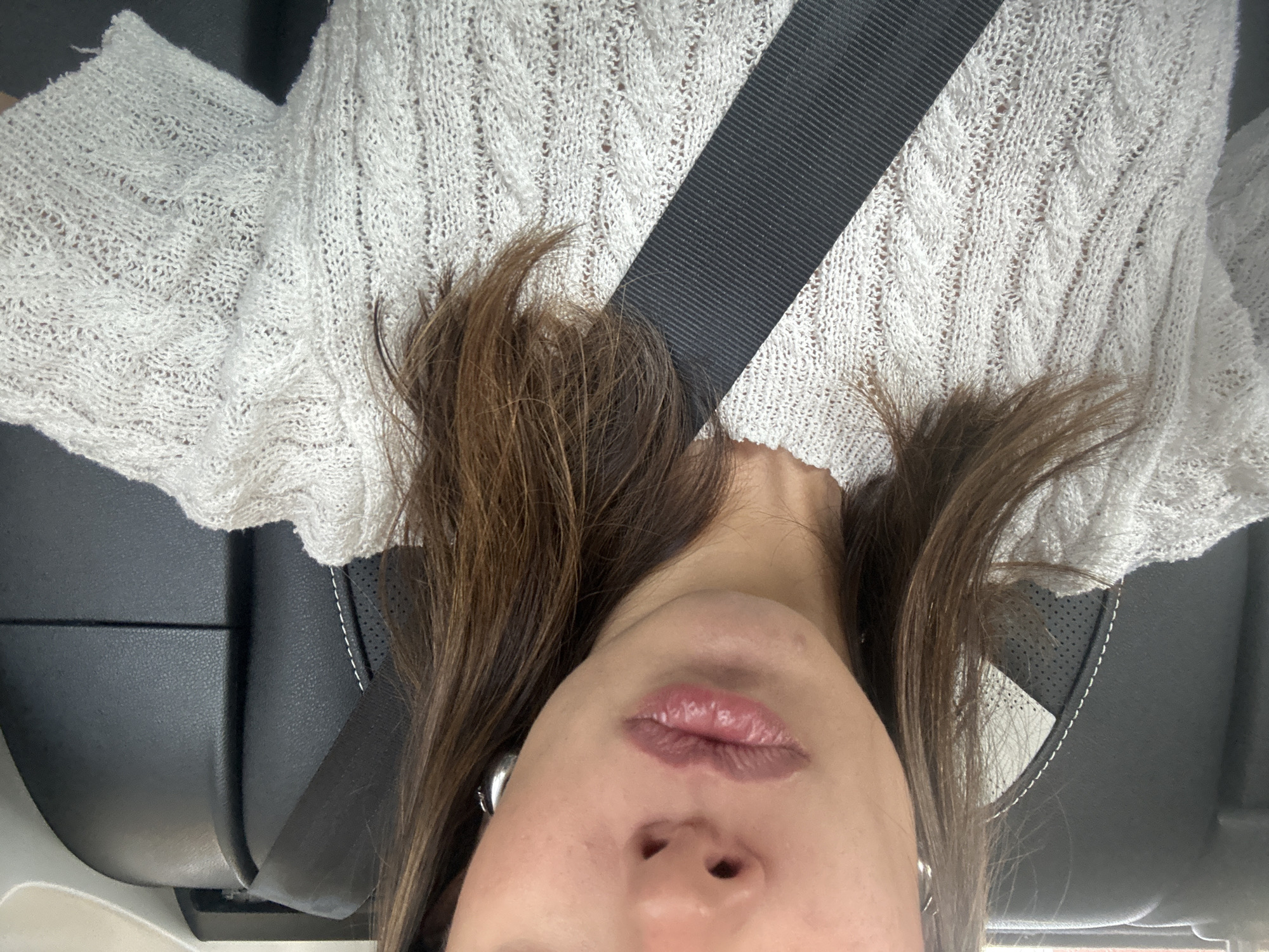 A close-up, upside-down perspective image shows a person in a vehicle wearing a seatbelt. The person has light brown hair and is wearing a white knitted top. The seat is black leather with white stitching. The exact location in the city of Unknown, Unknown, is not discernible from the provided image.