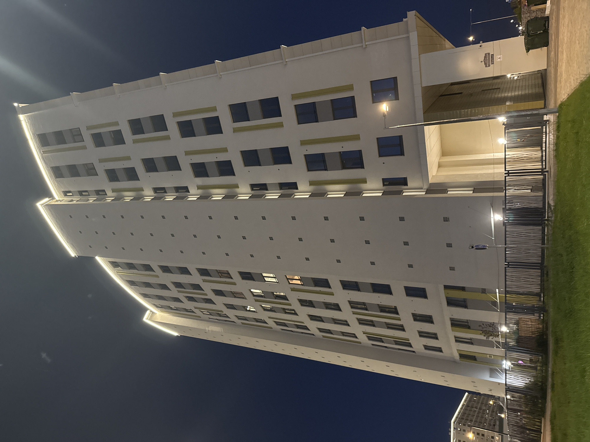 This is a nighttime, low-angle, wide shot of a modern, multi-story apartment building in Saint Petersburg, Russia. The building is predominantly white with vertical yellow accents and a series of uniformly spaced dark square elements on its facade. A string of warm-toned lights illuminates the rooftop. The sky is a deep, dark blue, indicating nighttime. In the foreground, there is a green grassy area and a black metal fence. To the left and further in the background, another building with illuminated windows is visible. A streetlamp with a warm light is positioned to the right of the main building. The scene appears to be a residential area.