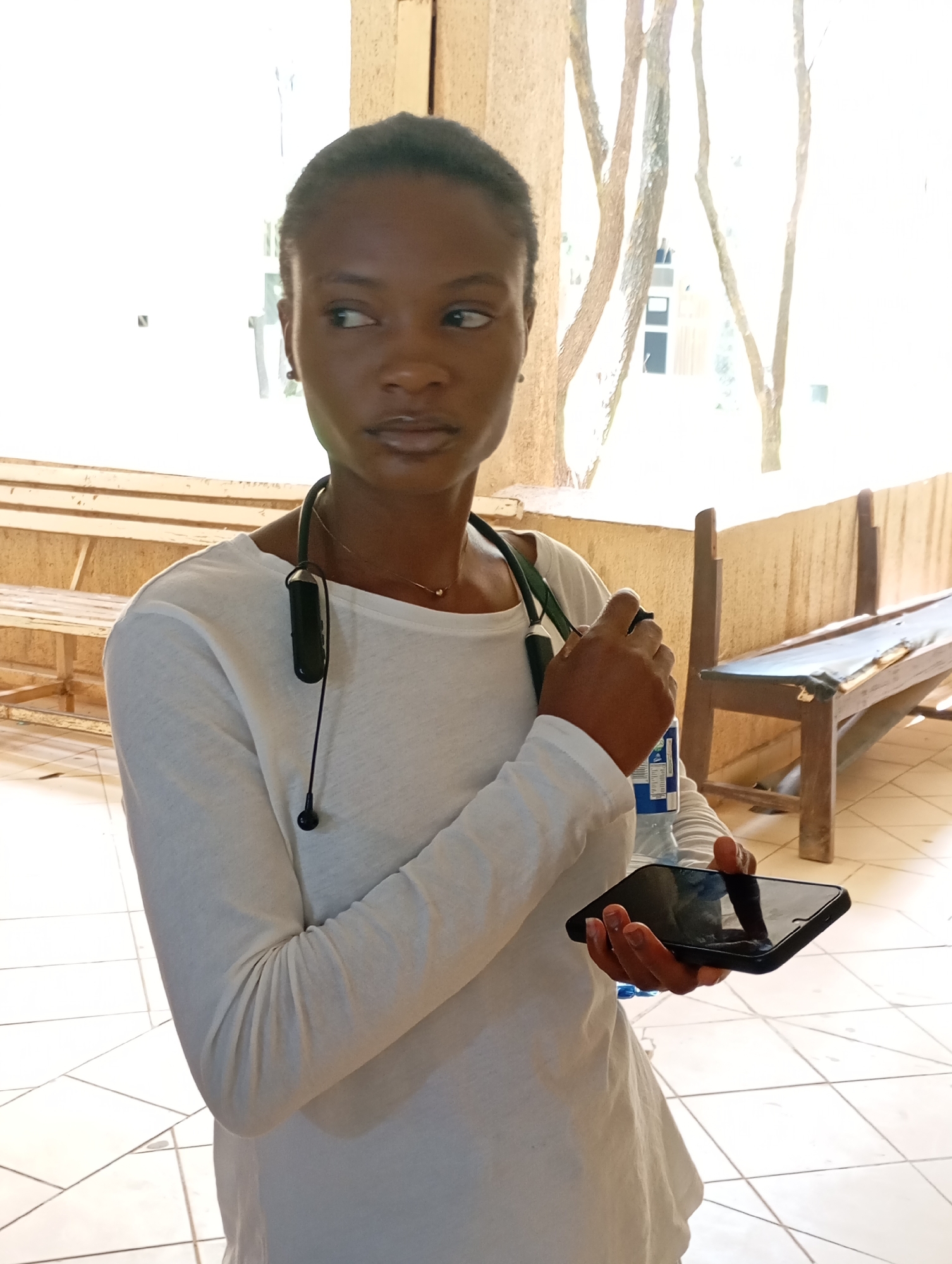 An African woman stands holding a black smartphone and a water bottle with a white label. She wears a white, long-sleeved shirt and dark wireless earbuds around her neck. The location appears to be a partially enclosed outdoor waiting area in Gwong, Nigeria, with light-colored tile flooring. There are wooden benches visible in the background. The lighting is bright.