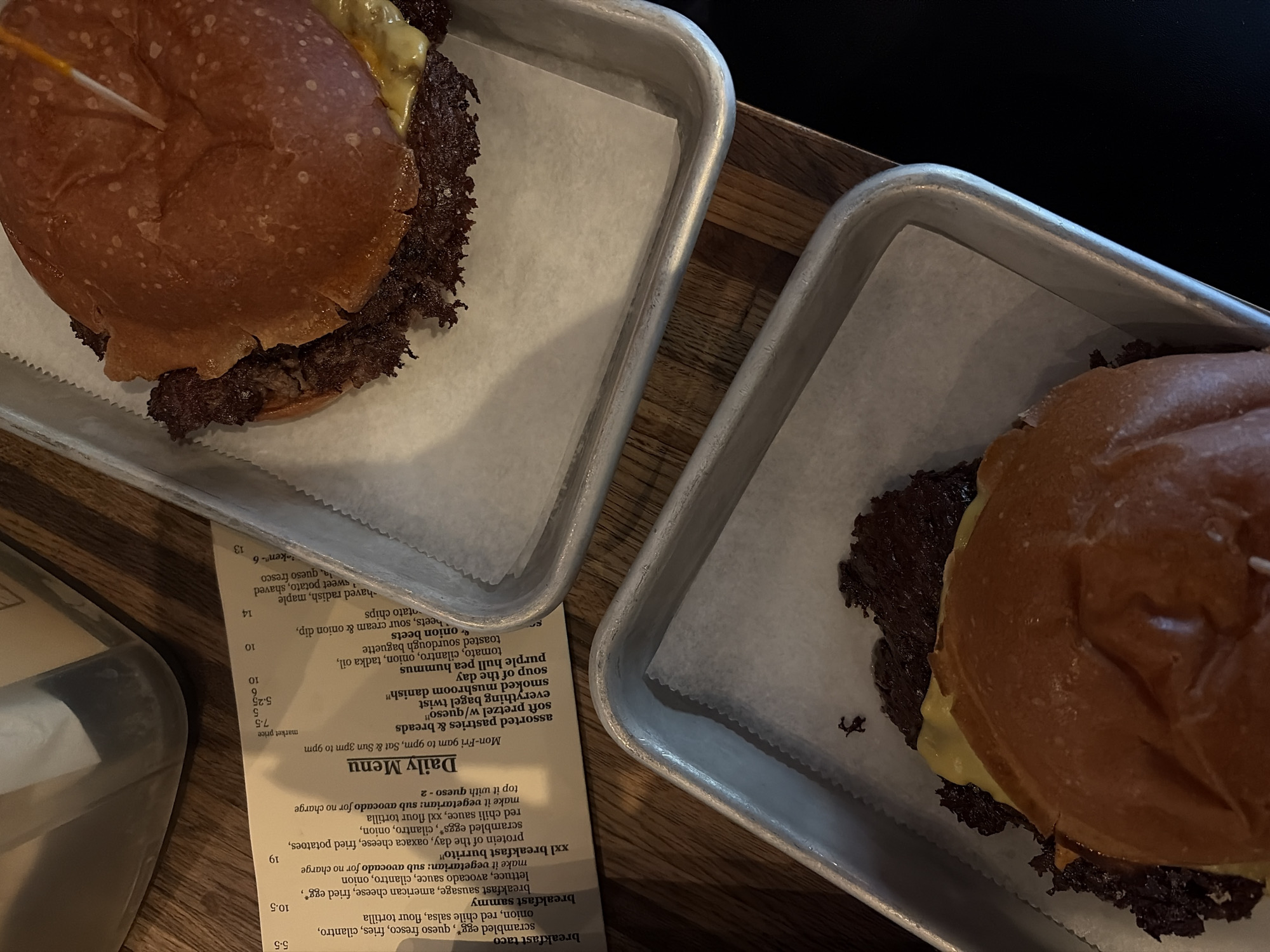 Two burgers, likely smash burgers, are presented in metal trays lined with parchment paper on a wooden table in a restaurant setting in Austin, United States of America. Both burgers feature a dark, crispy-edged patty with melted yellow cheese on a soft, browned bun. One burger has a toothpick inserted into its top bun. The scene suggests a meal about to be enjoyed.
A crumpled paper menu, titled "Daily Menu," is visible between the two trays. The menu indicates operating hours: "Mon-Fri 9am to 9pm, Sat & Sun 3pm to 9pm." Various breakfast and daily items are listed, including "breakfast taco," "breakfast sammy," and "xxl breakfast burrito," with ingredients such as "scrambled egg*", "queso fresco," "avocado sauce," and "red chile salsa." Other items include "assorted pastries & breads," "soft pretzel w/queso," "everything bagel twist," "smoked mushroom danish," "soup of the day," and "purple hull pea hummus." Prices ranging from 5.5 to 19 dollars are printed next to the items, with some marked "market price." The lighting in the image is dim, suggesting an indoor setting, possibly an evening meal or a dimly lit cafe.
