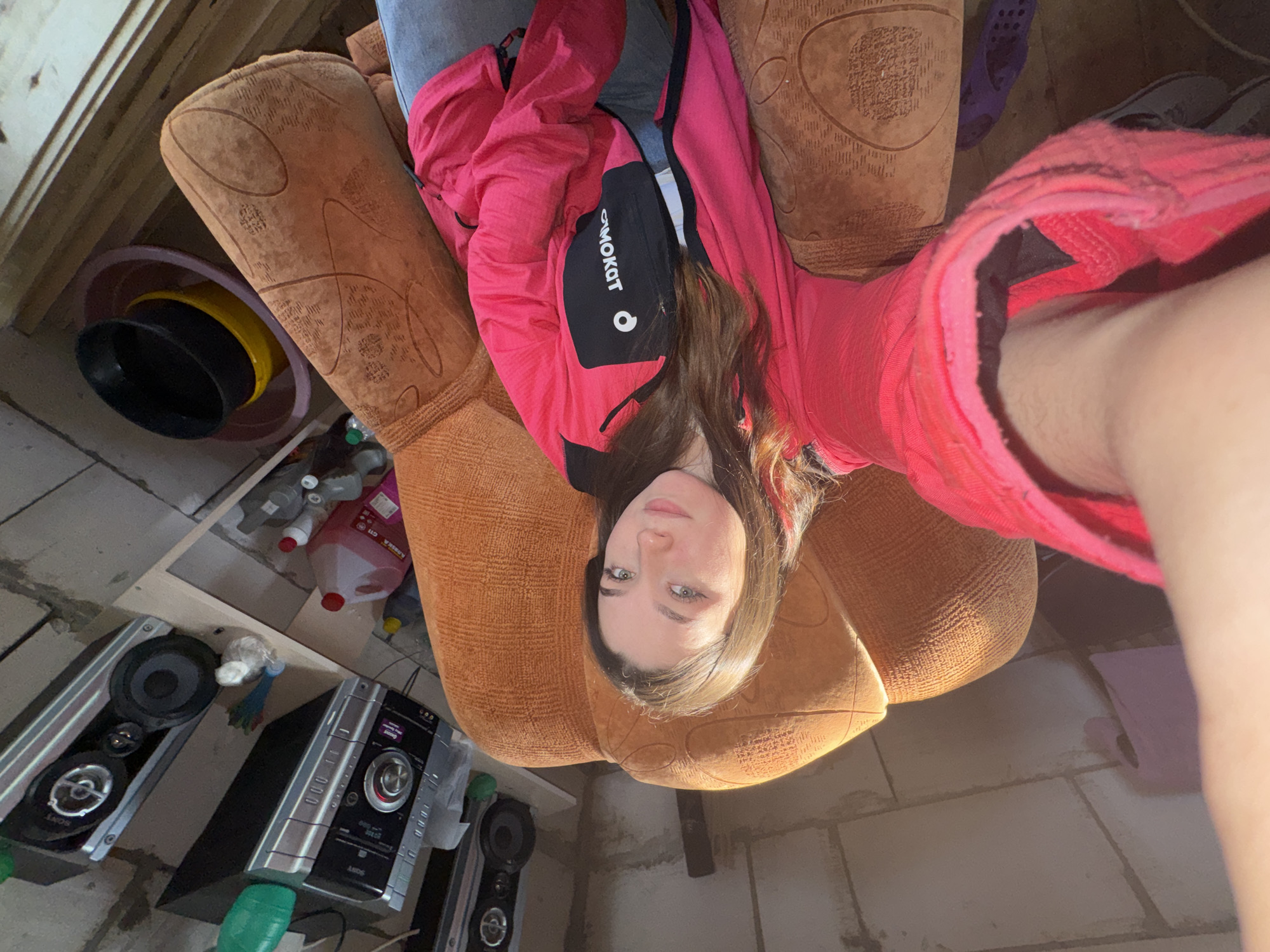 A forensic description of the image, depicting a scene in Krasnokamsk, Russia, is as follows:
A young adult female is visible in a selfie-style photograph, lying on an orange-brown upholstered couch with a patterned fabric. Her head is positioned towards the upper center-right, and her body extends diagonally towards the lower left. She has medium-length brown hair and light-colored eyes, looking directly into the camera. She is wearing a bright pink long-sleeved jacket with black accents, including a prominent black panel on the left chest that displays a reversed white logo, possibly "SIMOKAT". Underneath the jacket, a grey garment, likely pants, is visible. Her right arm, holding the camera, is extended towards the foreground, with a partial view of a pink sleeve.
The background reveals an indoor environment with utilitarian or unfinished aesthetics. To the left of the woman, on a wooden shelf, a silver-colored Sony stereo system is present, consisting of a central unit and two detached speakers with visible cones. A green plastic bottle is partially visible near the stereo. The wall behind this area appears to be constructed of light-colored concrete blocks or rough plaster.
To the right of the woman, further in the background, a stack of plastic basins and buckets is visible on a lower surface: a large, light purple basin at the bottom, holding a yellow bucket, which in turn holds a black bucket partially filled with liquid. Several other plastic bottles, some containing liquids, are on an adjacent shelf or floor. The wall in this section is also rudimentary, possibly wood paneling or unfinished masonry.
The floor is a light grey, possibly concrete or large tiles, with visible grout lines. Natural light, likely from an unseen window to the right, casts distinct shadows across the couch and the room. In the bottom right corner, a pair of dark blue/purple, perforated slip-on shoes are visible on the floor.