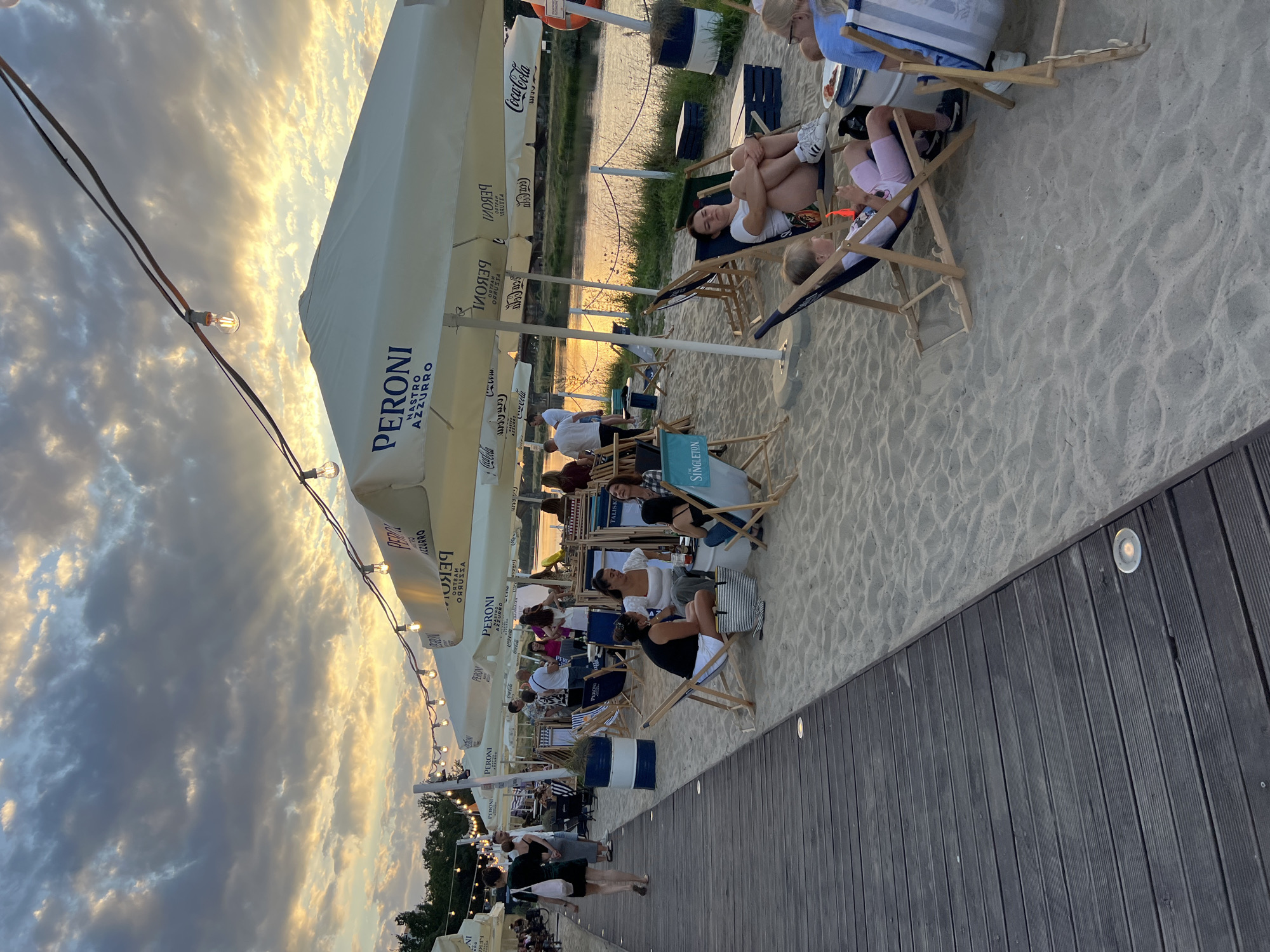 The image depicts a waterfront scene with a sandy area filled with beach chairs and umbrellas, characteristic of a summer recreational spot. People are seen relaxing, socializing, and enjoying the outdoors. String lights are visible overhead, suggesting an evening or late afternoon setting. The water body in the background, likely a river, reflects the warm hues of the sky, indicating a sunset or sunrise. In the foreground, a wooden boardwalk runs alongside the sandy area, providing a pathway. The overall ambiance is casual and leisurely, typical of a beachside bar or cafe. This scene is likely located at the Wrocław Beach, a popular recreational area situated along the Odra River in Wrocław, Poland.