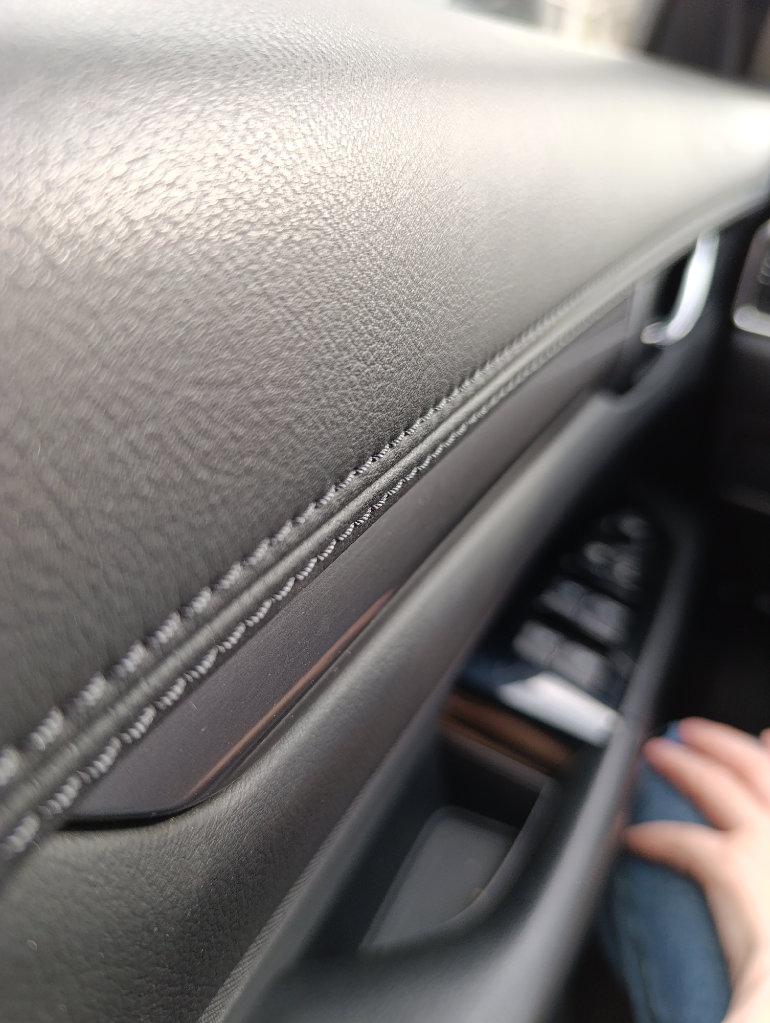 The media file provides a close-up, angled view of a car's interior, likely focusing on a door panel or dashboard section. The primary subject is a dark, textured material, resembling faux leather or a similar synthetic, which covers a significant portion of the surface. This material features distinct double stitching, executed in a lighter-colored thread, running horizontally across the panel.
Below the stitched line, a smooth, dark trim piece is visible, contrasting with the textured material. Further into the background, the image becomes increasingly blurred due to a shallow depth of field. Visible elements include a metallic door handle, a control panel with several indistinct buttons (likely for windows or other functions), and a storage compartment or door pocket.
In the lower right corner of the main image, a portion of a human hand, possibly a right hand, is visible, resting on what appears to be a leg clad in blue denim jeans. This indicates a person is present inside the vehicle. The lighting is bright and diffuse, suggesting it is daytime, possibly with an overcast sky or the car is parked in a shaded area, creating soft reflections on the textured surfaces. There is no visible text within the image. The scene is set inside a car in Chaikovsky, Russia.