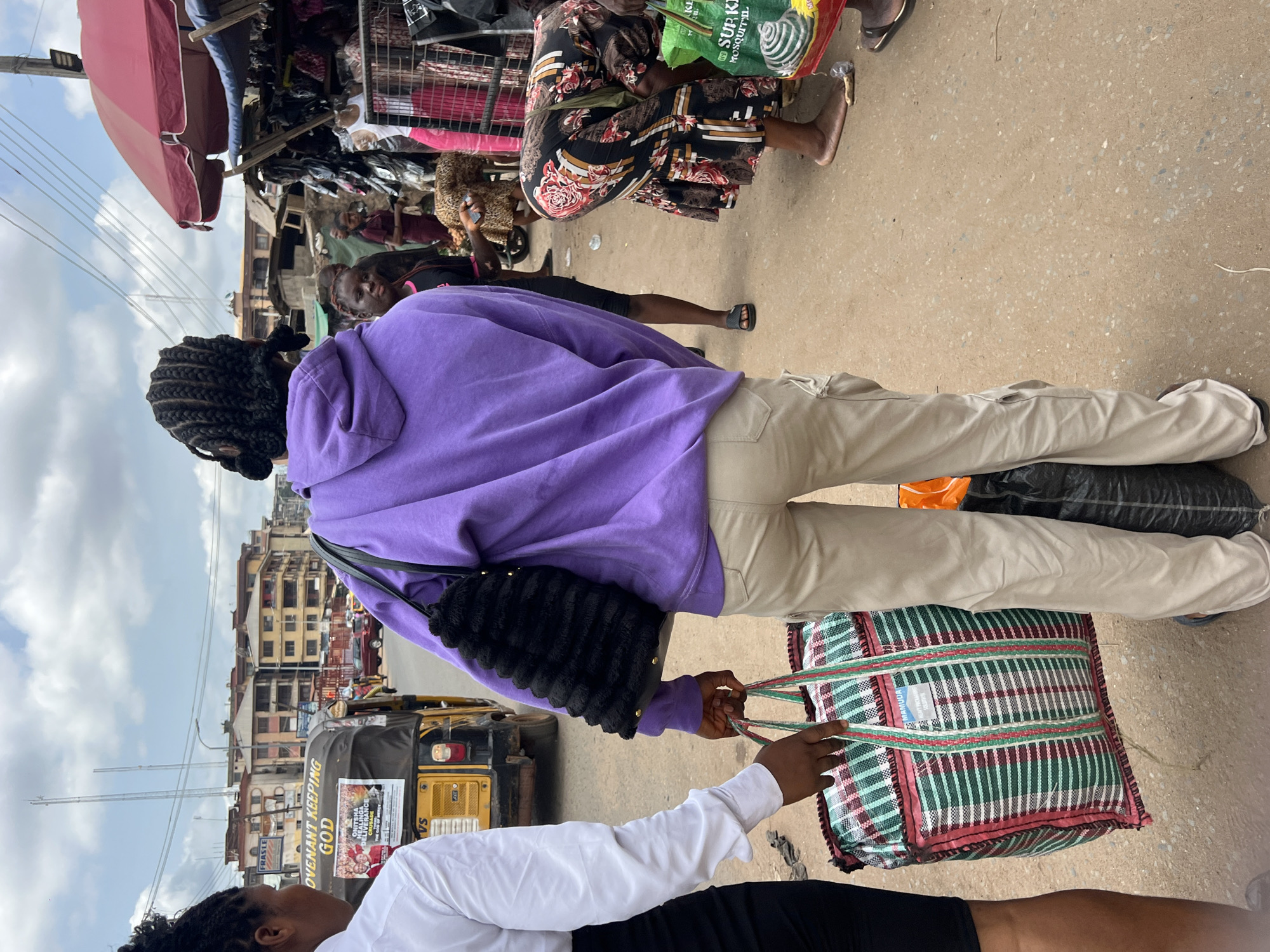 This outdoor image, taken in Nkpor, Nigeria, captures a vibrant street market scene under a cloudy daytime sky. The perspective is from behind and slightly above two individuals in the foreground, creating a sense of being part of the crowd.
The primary subject is a person facing away from the camera, wearing a purple hooded sweatshirt, light beige cargo pants, and carrying a black, textured handbag on their shoulder. They are holding the handle of a large, rectangular, woven bag with green, red, and white plaid patterns. Another person, seen partially in the lower left, wearing a white long-sleeved shirt and black skirt, is also holding the same patterned bag, suggesting they are sharing the load or exchanging the item. This large bag has visible text "MARHUDA" and "QUALITY BAG".
In the middle ground and background, a bustling market environment unfolds. Several market stalls are visible, laden with various goods, including clothing and other items. A vendor wearing a patterned dress is seated near one of these stalls, and another person holds a green plastic bag with partially visible text "SUP KIL MOSQUITU" (likely for mosquito repellent). People are scattered throughout the scene, walking and interacting.
The background reveals a street with older multi-story buildings, characteristic of an urban area. A yellow motorized tricycle, commonly known as a Keke Napep, is parked on the left, displaying religious text such as "COVENANT KEEPING GOD" and "ORIGINAL HEALING AND DELIVERANCE CRUSADE MARANATHA PROPHECY MINISTRY." Overhead, power lines crisscross the sky. The ground is a light-colored paved surface, typical of roads or walkways. The overall atmosphere suggests a busy, everyday commercial activity.