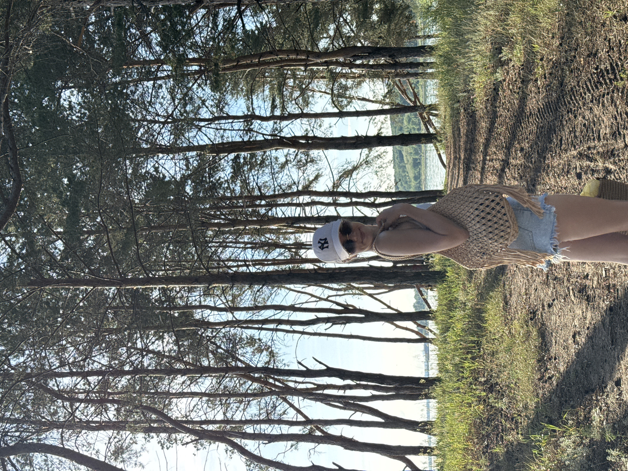 A young woman stands on a path in a pine forest near a body of water. She is wearing a white baseball cap, sunglasses, a light-colored top, and denim shorts. She is carrying a woven bag over her shoulder. The path is dirt and has tire tracks. The trees are tall and thin, with a dense canopy. The sky is blue with some clouds. The sun is shining, casting shadows on the path. In the background, there is a body of water and a distant shoreline with trees.
