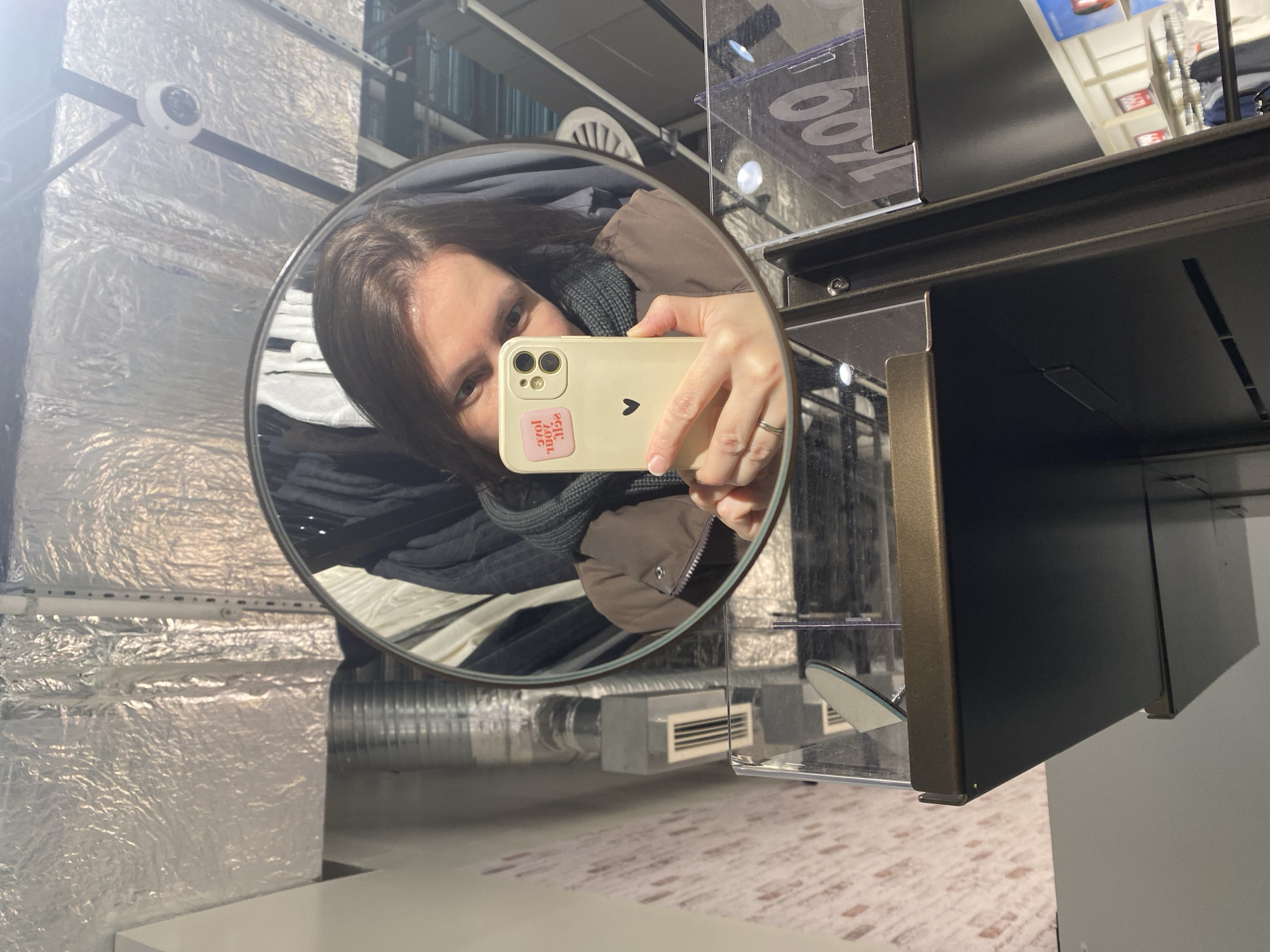 A woman is taking a selfie in a round mirror. She is wearing a brown jacket and a grey scarf. The phone she is holding is beige and has a pink sticker. The mirror is attached to a dark brown metal structure. The background shows a silver duct, a security camera, and clothes on a rack. The location is likely a store, possibly in Yoshkar-Ola, Russia, but there is no verifiable information to confirm this location in the image.
