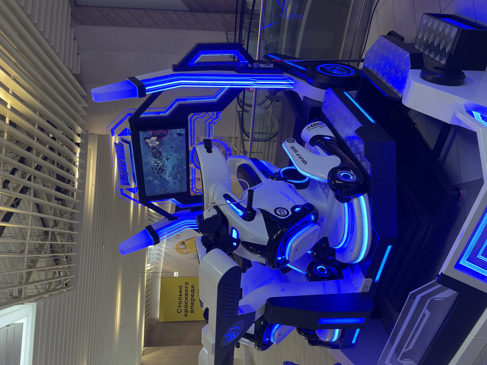 This is a high-angle, close-up shot of a futuristic-looking arcade game machine in Kazan, Russia. The machine features white and black components with blue LED lighting. It includes a central screen displaying a game interface. The design incorporates simulated seating and control elements, suggesting a virtual reality or immersive gaming experience. In the background, a section of the building's interior is visible, including a ceiling structure and a sign written in Russian.