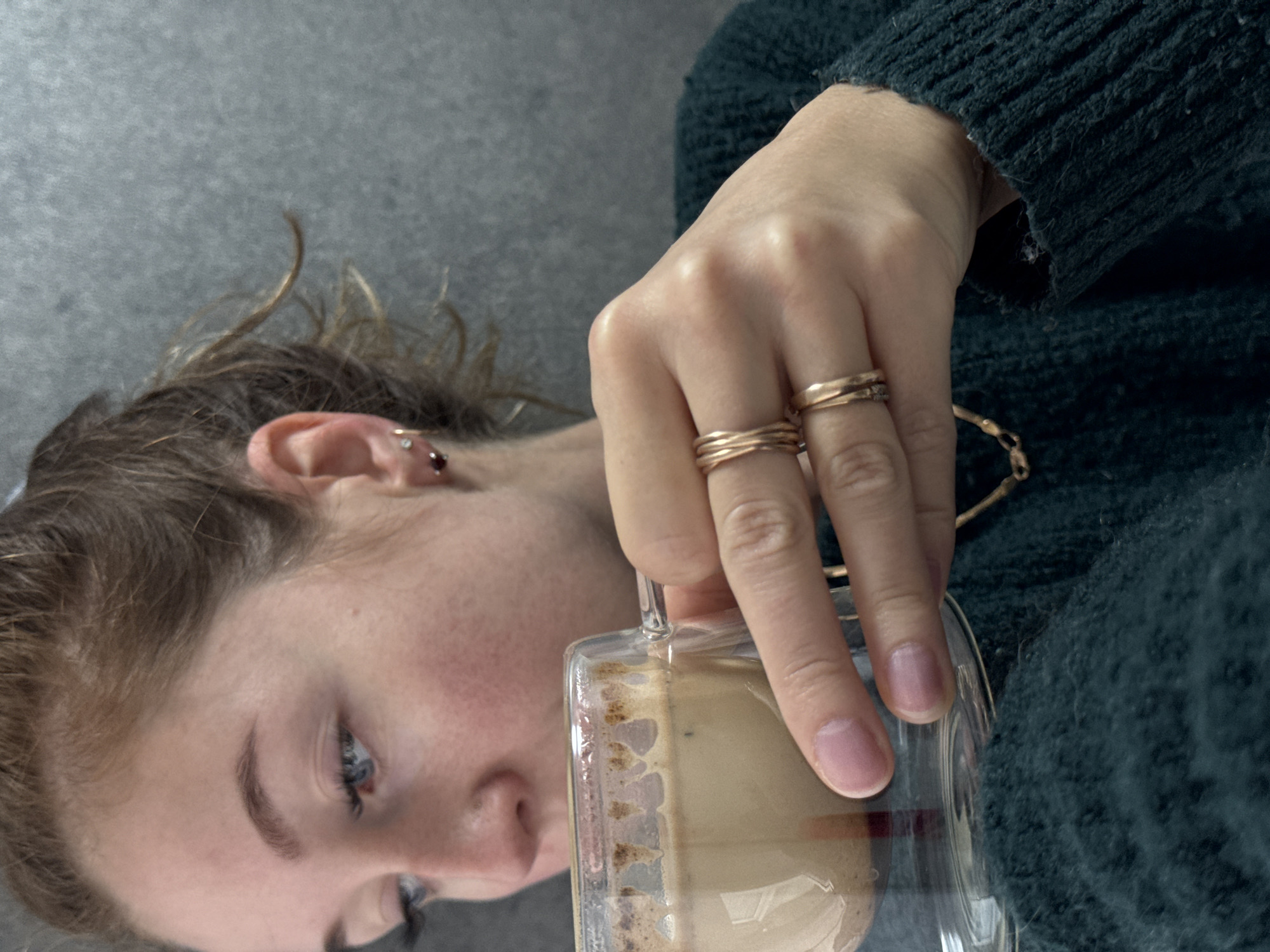A young woman with fair skin and blue eyes, wearing a dark green sweater and gold rings, is holding a glass mug filled with a light-colored beverage, possibly coffee or a latte. The woman's face is angled slightly downward and to the left. Part of her ear is visible, showing multiple piercings. The background is a neutral grey. There is no evidence of the location being Chaikovsky, Russia.
