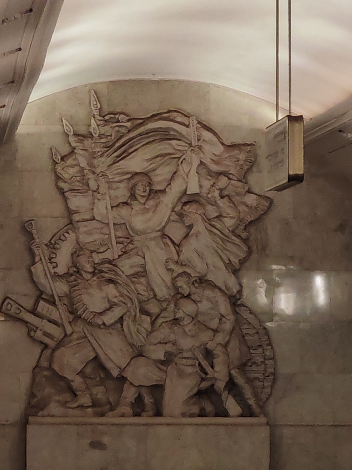 A large bas-relief sculpture, carved into a polished stone wall, depicts a dynamic and heroic scene. The sculpture features multiple human figures, primarily male, rendered in a style characteristic of Socialist Realism.
At the center-top, a prominent figure, likely a soldier or revolutionary, stands with one arm raised high, holding a large flag or banner that billows behind them. Other figures surround this central leader, some appearing to be soldiers armed with rifles and spear-like weapons. One figure on the lower left is shown with what appears to be a rifle and ammunition belts. Elements resembling a tank track or industrial gears are visible beneath some of the figures, suggesting a theme of military might or collective labor.
The overall composition conveys a sense of movement, struggle, and determination, possibly depicting an advance, a revolutionary uprising, or a moment of triumph. The figures are clad in uniform-like attire.
The sculpture itself is monochromatic, executed in the same light, earthy tone as the surrounding polished stone wall, which appears to be marble or a similar material. The setting is indoors, featuring an arched ceiling and a rectangular, metallic light fixture suspended from above on the right side of the frame. Reflections from an unseen light source are visible on the polished wall.
There is no visible text on the sculpture or the wall. The style and context suggest this is a monument within a public space, possibly a metro station, consistent with the given location of Moscow, Russia.
