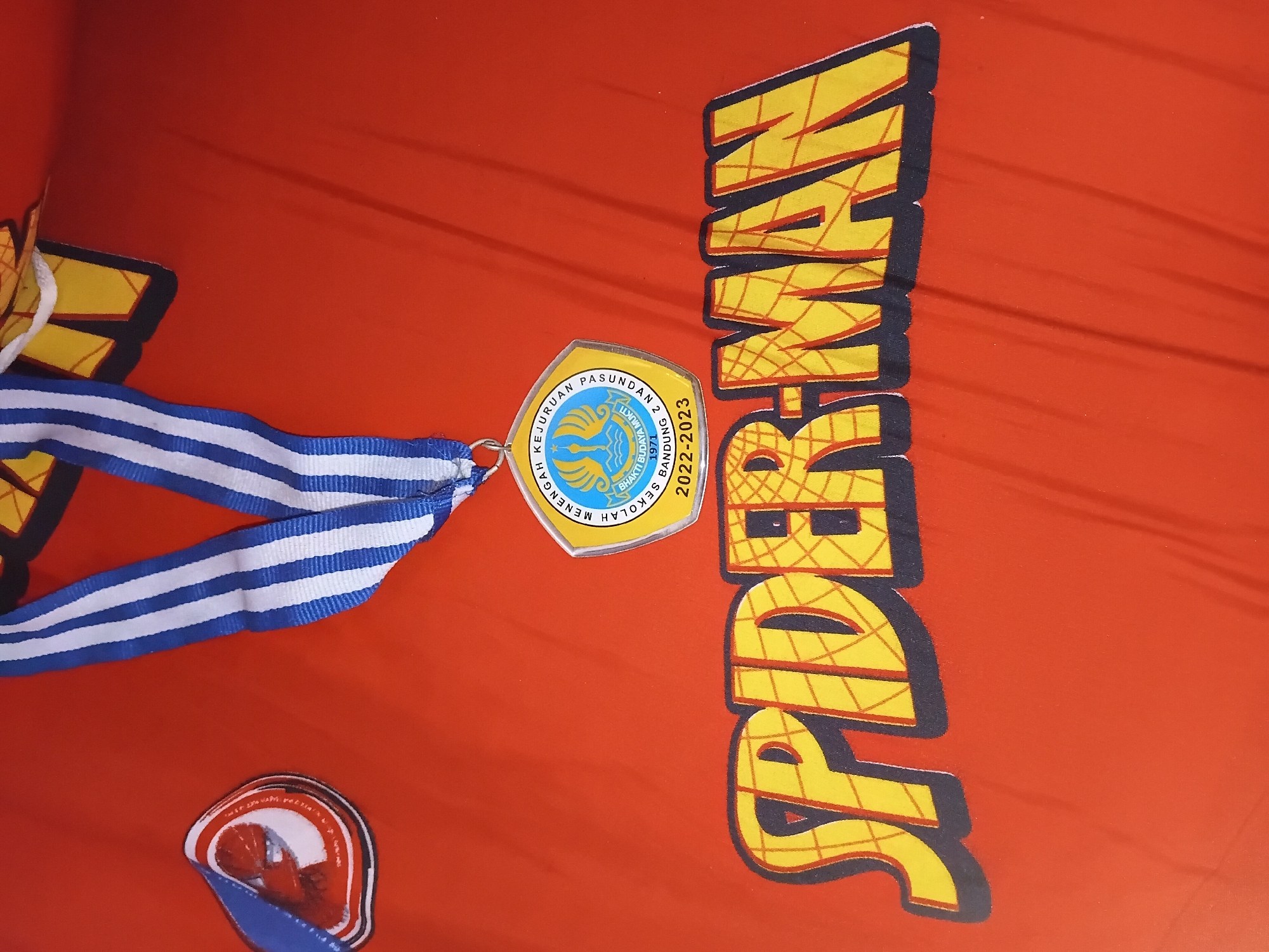 The image displays a close-up, overhead view of a red surface, likely fabric, with several objects. Prominently featured is the word "SPIDER-MAN" in large, bold, yellow letters with a dark blue outline, mimicking the iconic comic book lettering. Above the text, a medal hangs from a blue and white striped ribbon. The medal itself is hexagonal with a yellow border and contains a blue and yellow emblem with text. The emblem includes "SEKOLAH MENENGAH KEJURUAN" (Vocational High School) in blue, and within the emblem, text reads "BHAKTI BUDAYA MKU" and "1971 BANDUNG 2". Below this text, it says "2022-2023". To the left of the medal, a small, circular sticker with a red and white design featuring a character's face, possibly Spider-Man, is partially visible. The lighting suggests an indoor setting, and the red surface appears to be a piece of clothing or bedding. The presence of the school name and "Bandung" on the medal indicates a connection to Bandung City, Indonesia.