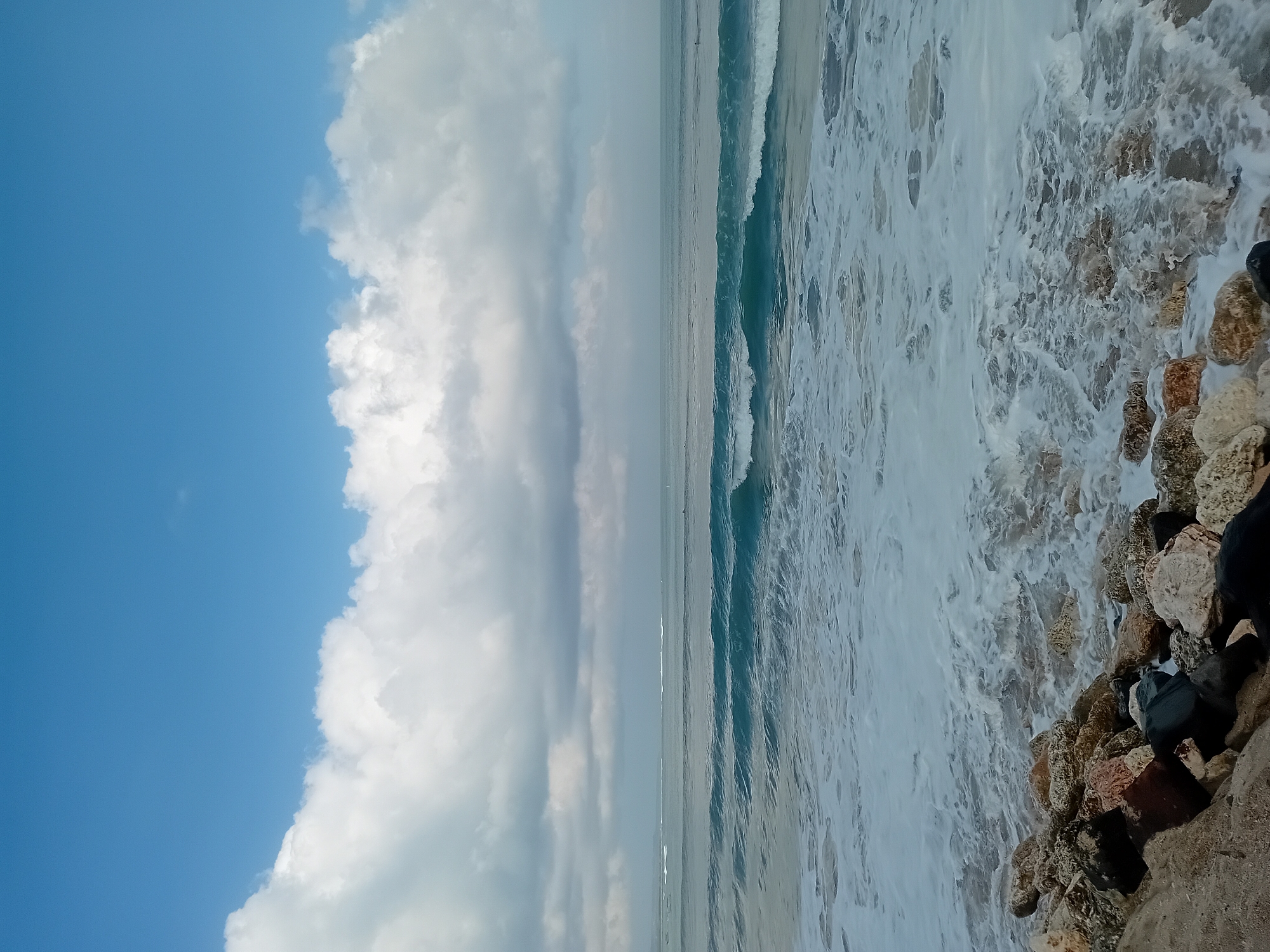 The image depicts a serene seascape with a rocky shore in the foreground, waves crashing against the rocks, and the vast ocean stretching out to the horizon under a cloudy blue sky. The location is identified as the city of Kuta, Indonesia, a well-known tourist destination famous for its beaches and surf. The scene is a natural one, devoid of any specific human activities or interactions, presenting a peaceful and tranquil view of the ocean. The photograph captures the essence of a tropical beach, with the clear blue sky and white clouds contrasting with the turquoise hues of the ocean waves. The rocky shore adds a rugged texture to the scene, enhancing its natural beauty. The overall impression is one of a serene and picturesque coastal landscape.