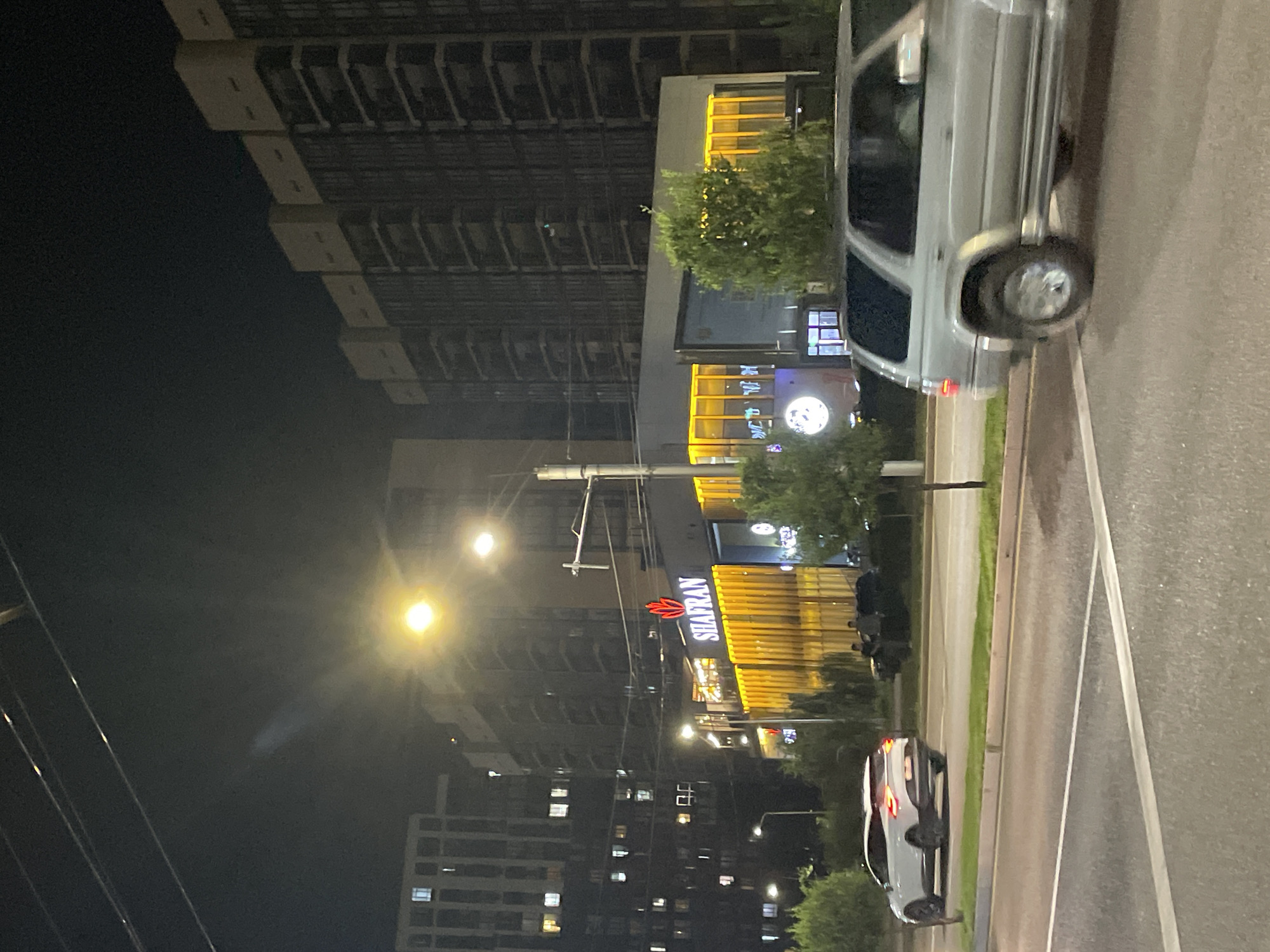 The image captures a nighttime street scene in Almaty, Kazakhstan, featuring modern architecture and traffic. In the foreground, a silver SUV is driving on the right side of a multi-lane road. The road surface appears to be asphalt with white lane markings. To the left of the road, a white car is parked, facing away from the camera.
Behind the road, a contemporary building with illuminated signage that reads "SHAFRAN" stands out. The building has a modern facade with golden-colored vertical elements and large windows. Trees are planted along the sidewalk in front of the building.
In the background, several tall residential buildings with multiple floors and illuminated windows dominate the skyline. The overall ambiance is that of a well-lit urban environment at night.