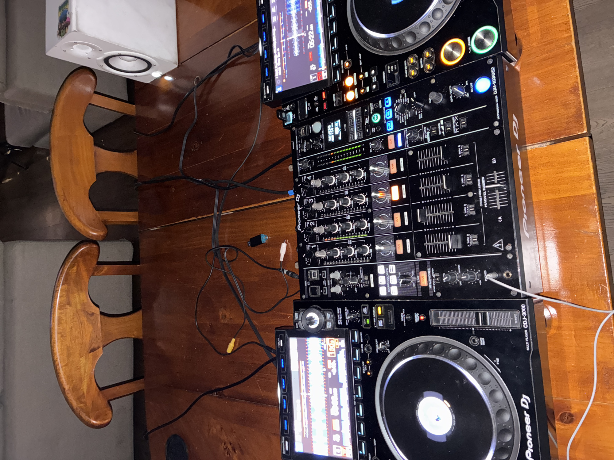 The image shows a Pioneer DJ setup, consisting of two CDJ-3000 multi-players and a DJM-900NXS2 mixer, placed on a wooden table. To the left of the setup, there is a white speaker and two wooden chairs. The image is a close-up shot, focusing on the DJ equipment and its arrangement on the table. The location is in Mexico City, Mexico.