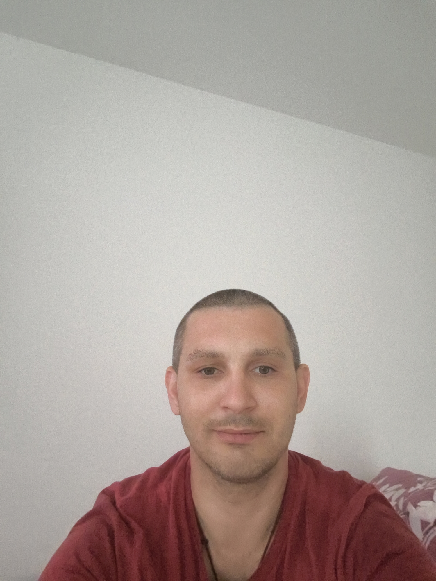 A selfie of a man with a buzz cut and a light beard is shown. He is wearing a maroon t-shirt. He is looking at the camera with a neutral expression. The background is a plain, light-colored wall. The image appears to have been taken indoors. There is no visible indication of the city of Wolgast, Germany.