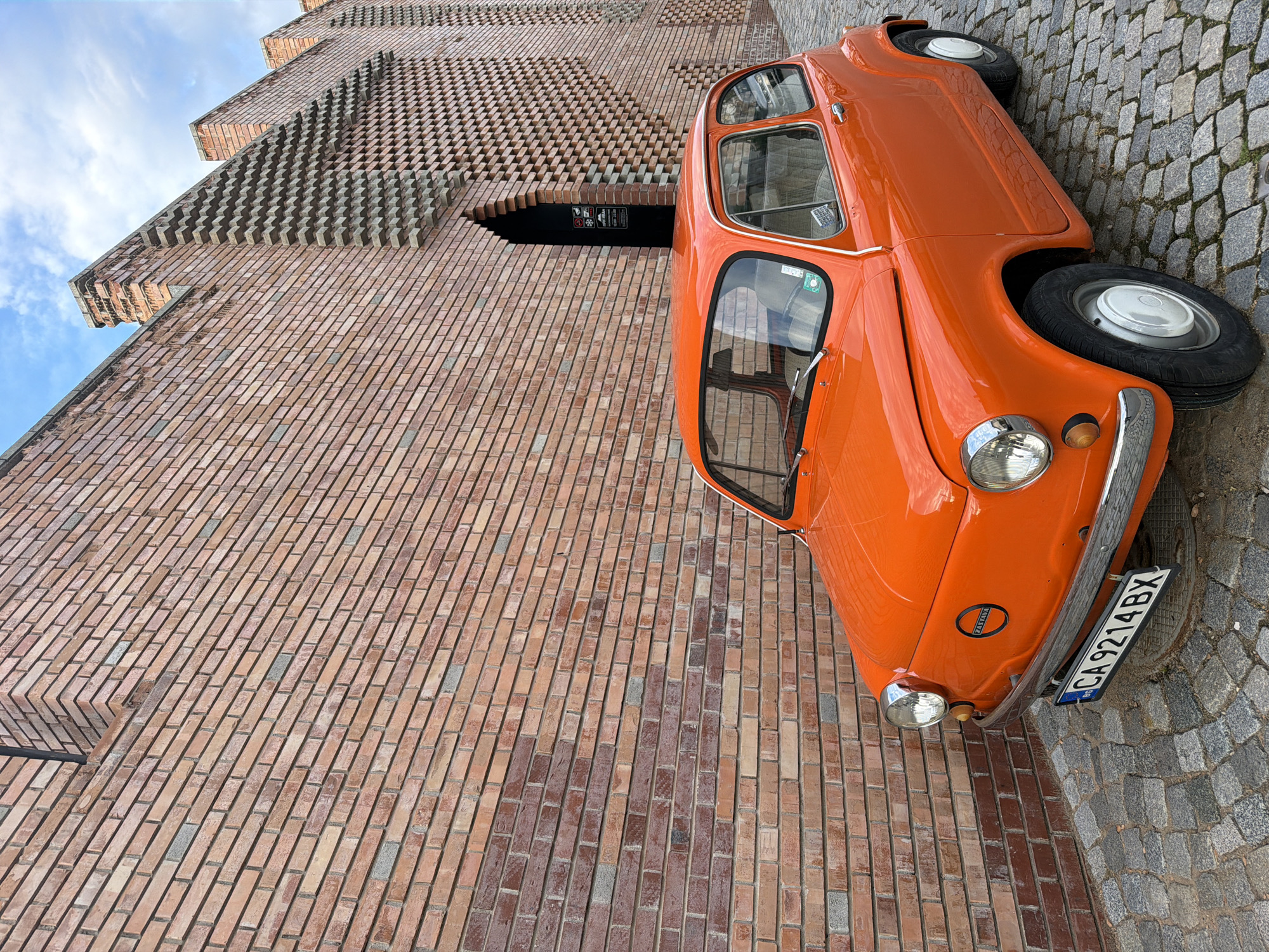 The image shows an orange Zastava 750 car parked on a cobblestone road next to a brick building in an urban setting. The building features a brick facade with a protruding layered design near the top, and a dark arched entrance. The car's license plate reads "CA 9214 BX". The sky is visible in the upper left corner, with some clouds. The city and country are unknown.