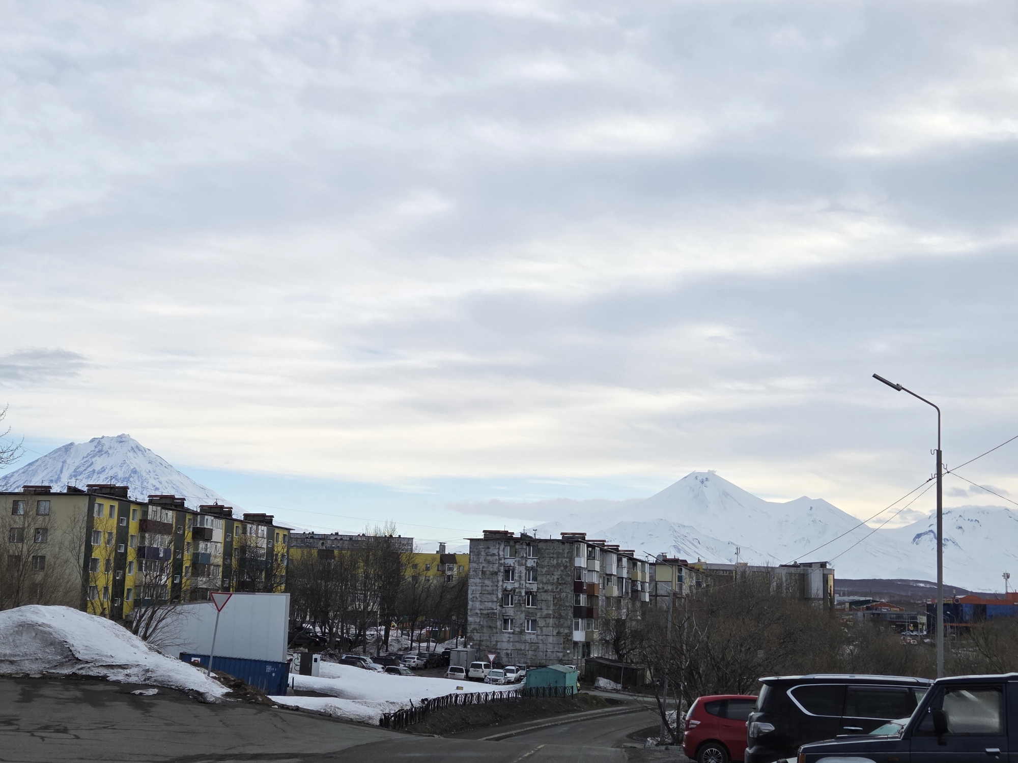 This photograph captures a city landscape in an unknown location, possibly in early spring based on the remaining snow. The scene is dominated by a cloudy sky, which covers most of the frame. In the midground, there are several Soviet-style apartment buildings in varying states of repair, some painted with yellow accents and others with drab concrete exteriors. Patches of snow remain on the ground, especially around the buildings and along the roadside. Several vehicles are parked in a lot behind the buildings and along the road, with a red hatchback and several dark-colored SUVs visible in the foreground. In the background, snow-capped mountains stand out against the skyline, adding a dramatic contrast to the urban setting. A streetlight is visible on the right side of the frame. The overall composition provides a stark view of urban life against a backdrop of natural beauty.