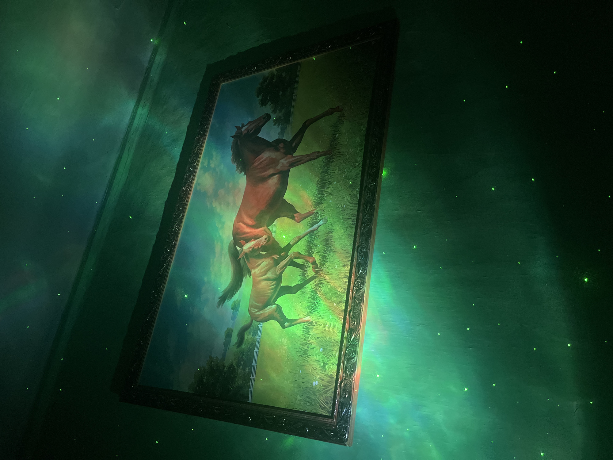 A framed painting of two horses running in a grassy field is illuminated by green ambient light that creates a surreal, almost cosmic effect on the surrounding walls. The painting, which features a light brown horse and a darker reddish-brown horse, is centrally placed, with the green light casting vibrant, swirling patterns across its surface and the wall. Small, bright green dots, resembling stars or fireflies, are scattered across the green-lit walls, enhancing the otherworldly atmosphere. The overall scene is dark, with the painting and the green light being the primary sources of illumination.