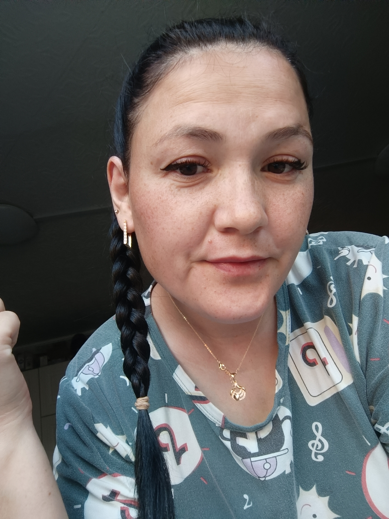 A medium close-up, selfie-style shot shows a woman with dark hair in a braid, wearing a patterned shirt and a gold necklace. She is looking directly at the camera with a neutral expression. The background is a dark, out-of-focus wall, suggesting an indoor setting. The image is well-lit, with the woman's face being the primary focus. There are no clear indications of the location being Karsha, Russia, or any other specific city.