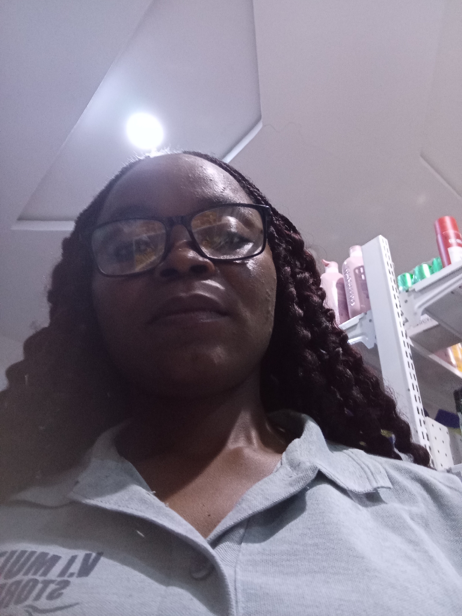 A woman with dark skin, wearing glasses and a light-colored collared shirt, is captured in a low-angle, indoor selfie. Her hair is styled in dark, long braids. She appears to be in a retail environment, with shelves of products visible in the background, suggesting a store setting. The lighting is bright, with a prominent light source above her. The text on her shirt is partially visible and appears to be in white lettering against the shirt's darker fabric, though the full text is not discernible. The environment suggests a commercial space in Howayi, Nigeria.