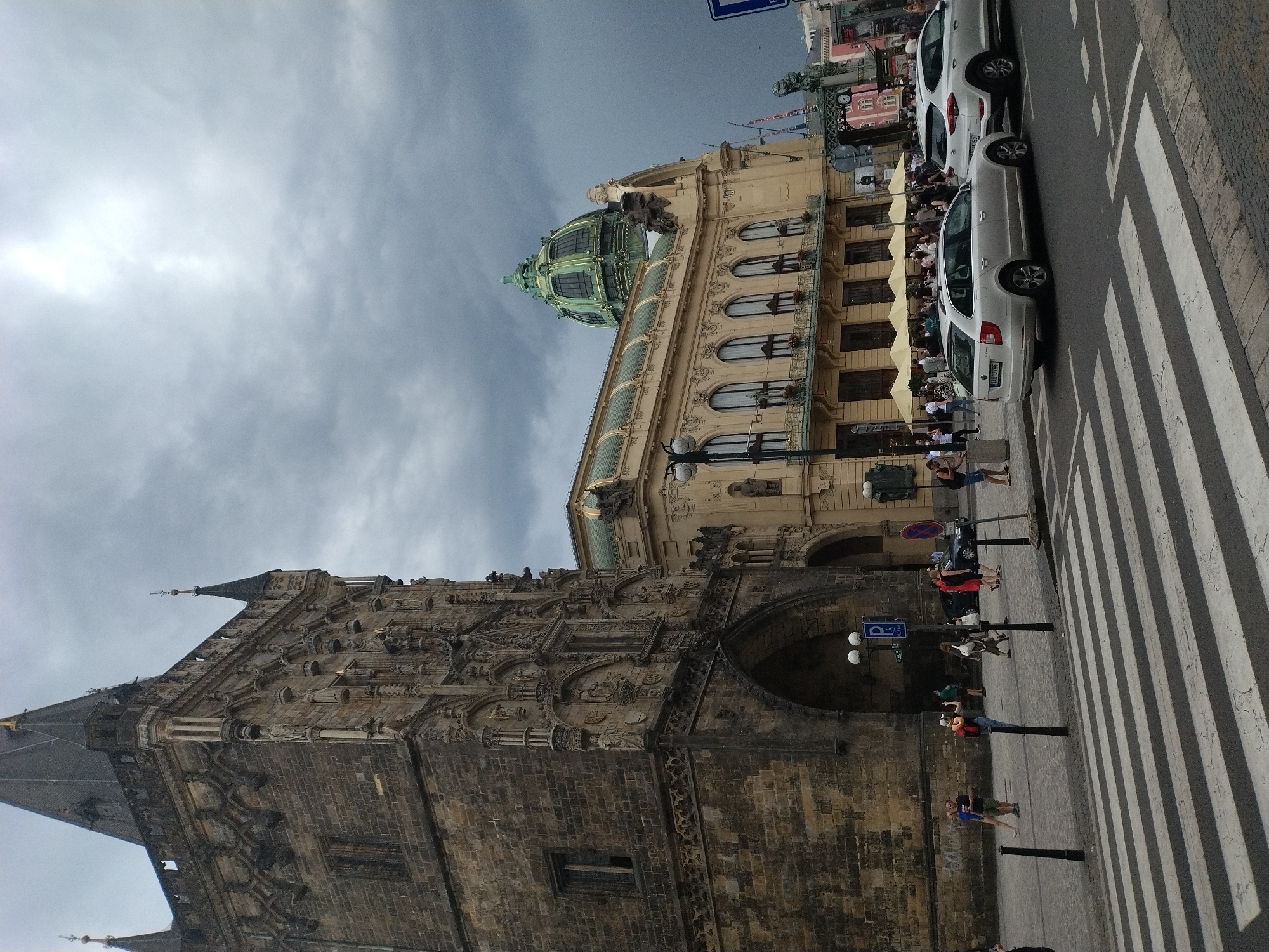 In the city of Prague, Czechia, an overcast sky looms over the Powder Tower, a gothic structure made of dark stone. To the right of the Powder Tower, a light-colored building with a green dome stands prominently. In the foreground, a pedestrian crosswalk spans a street, with several cars stopped at the intersection. Pedestrians are visible on the sidewalks, some walking and others gathered near the buildings. The scene captures a slice of urban life in Prague, with historic architecture set against a dramatic, cloudy sky.