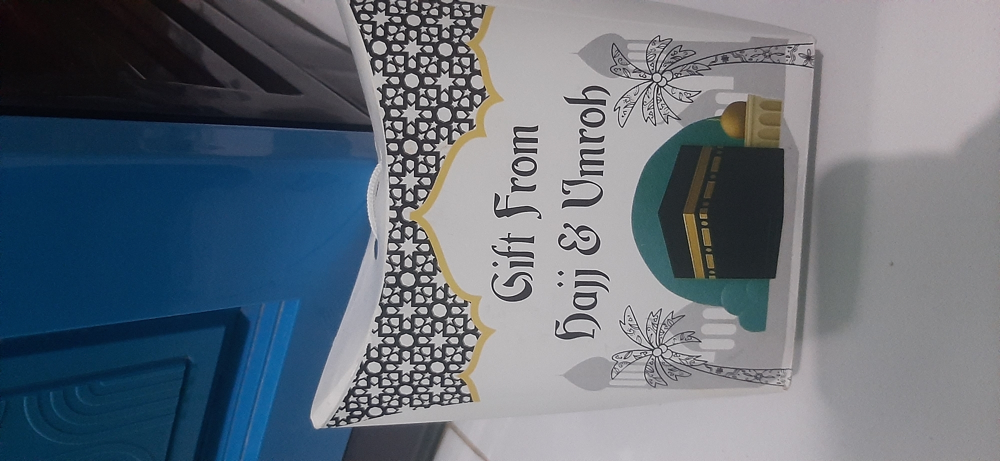 The image shows a white gift box with black and gold Islamic geometric patterns along the top edge. The front of the box features the text "Gift From Hajj & Umroh" in a dark, ornate font. Below the text, an illustration depicts the Kaaba in Mecca, surrounded by stylized palm trees and mosque-like structures. A small golden dome sits to the right of the Kaaba. The box is resting on a white surface, possibly a table, with a blurred blue object in the background. The lighting suggests an indoor setting.