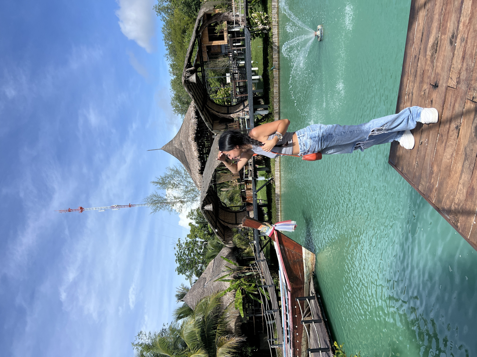 The image depicts an outdoor scene in Tambon Pa Kae Bo Hin, Thailand, featuring a woman standing on a wooden dock. She is wearing ripped light-wash jeans, a grey tank top, and white sneakers, and is holding an orange purse. She is shielding her eyes with her hand as she looks out over a body of turquoise water. In the background, several thatched-roof structures are visible, along with lush green vegetation and palm trees. A wooden boat with a red hull is docked to the left, and a fountain sprays water in the middle of the water. A tall, red and white communication tower stands in the distance against a blue sky with scattered clouds.