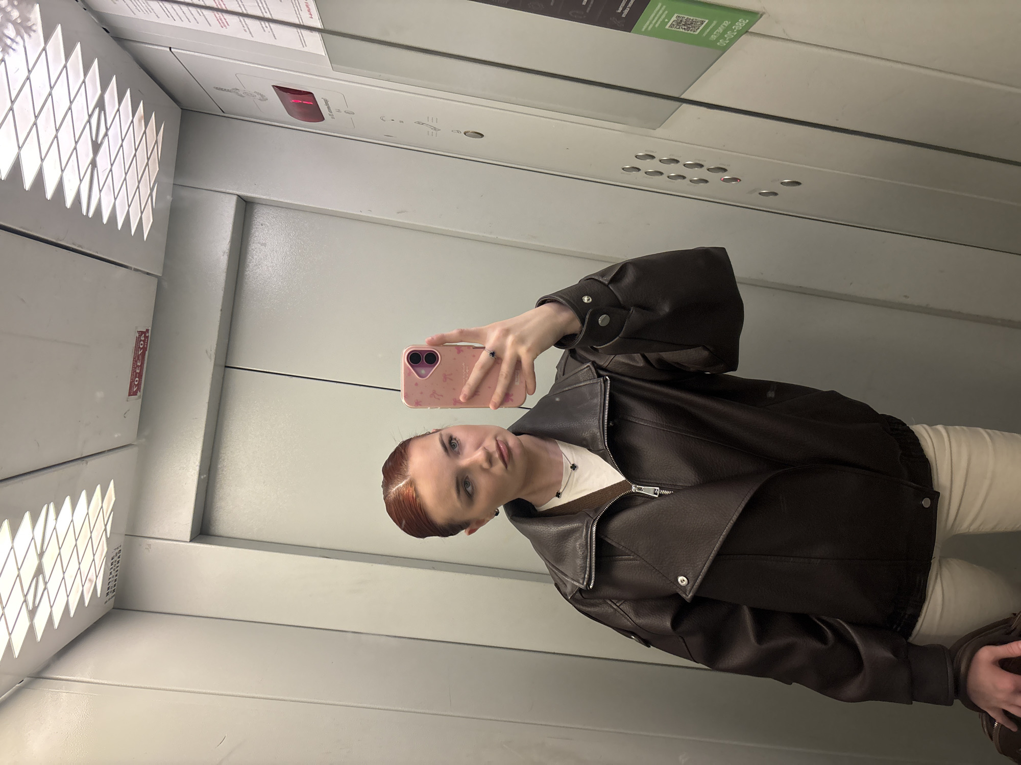 A young woman with red hair and subtle makeup takes a selfie in an elevator. She is wearing a brown leather jacket over a white top, and cream-colored pants. Her hair is pulled back from her face, with a distinct side part. She is holding a pink phone with a bunny case. The elevator walls are a light gray and have a metallic sheen. There is a diamond-patterned grate on the left side of the image, emitting light. Above her, a mirror reflects the elevator's control panel, which includes several buttons and a red illuminated display. A green QR code sticker is visible in the reflection, with some Russian text below it. The overall setting is an indoor elevator with a modern, utilitarian aesthetic. The woman's expression is neutral, and the lighting is bright, suggesting it is daytime.
