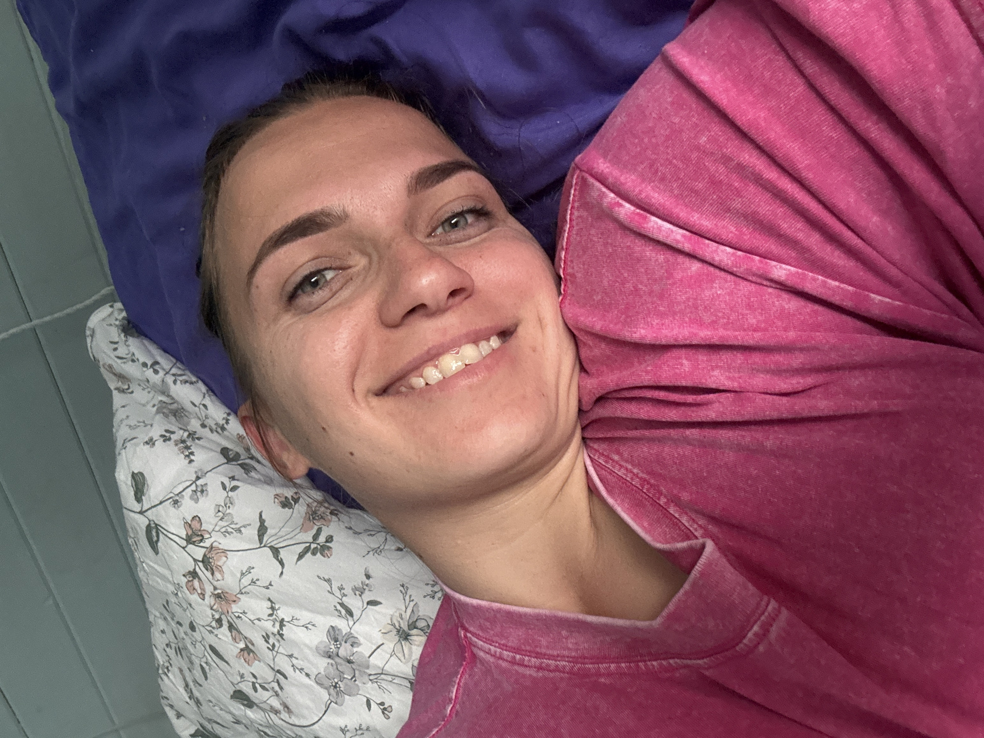 A person is lying on a bed with their head on a floral patterned pillow and their body angled towards the right. The person is wearing a pink shirt and smiling. A purple pillow is behind their head. The background includes what appears to be a wall. The image does not offer details to confirm the location in the city of Раздолье, Russia.