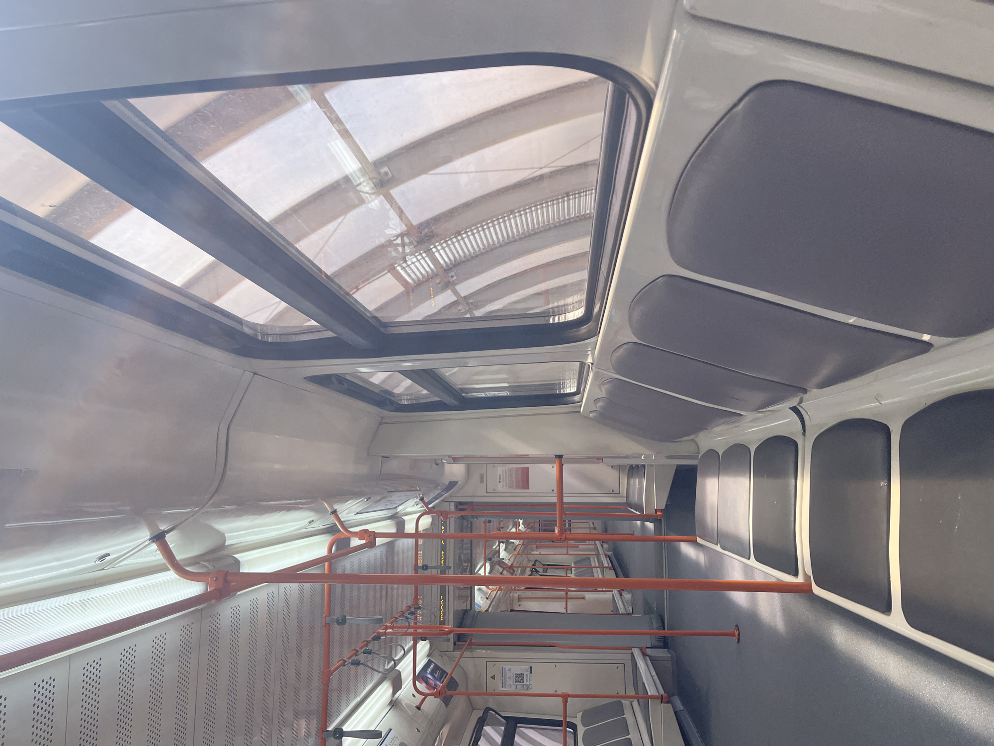 The image depicts the interior of a public transportation vehicle, likely a tram or light rail, in Sofia, Bulgaria. The vehicle appears empty. The interior features a white and gray color scheme with orange handrails. Overhead are skylight windows providing natural light. The seating arrangement is along the sides. The floor appears to be dark gray.