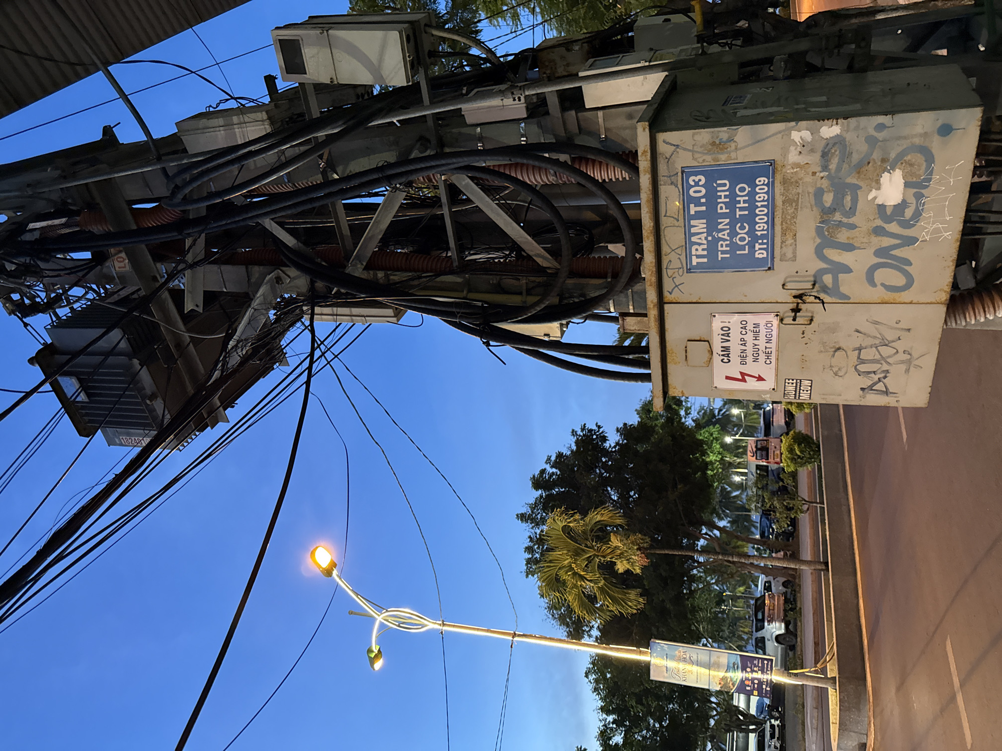 The image captures an electrical transformer pole and its complex network of wires against a twilight sky in Nha Trang, Vietnam. A street light with two illuminated lamps is visible on the left, casting a warm glow. The transformer unit itself is covered in graffiti, with the words "Trạm T.03 Trần Phú Lộc Thọ ĐT: 19001909" inscribed on a blue sign attached to it, indicating its location. To the left of the pole, a banner with text in Vietnamese, likely an advertisement or informational poster, is also present. The background shows a line of trees, palm trees, and parked vehicles, suggesting a street or roadside setting. The overall scene depicts a common urban utility infrastructure in Vietnam.