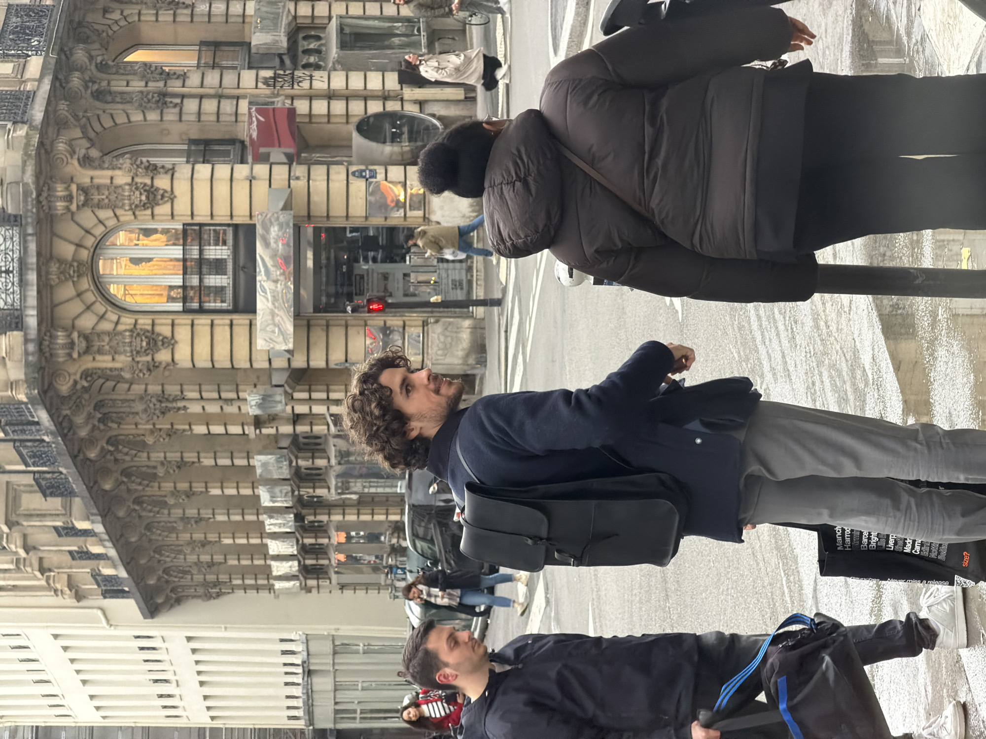 The image depicts a street scene in Paris, France. The camera angle is vertical. Two buildings with ornate architectural details are visible. Pedestrians are present in the foreground, including a man with curly brown hair wearing a navy blazer and gray pants, and carrying a black bag. Another man wearing black clothing and carrying a bag with blue straps is also in the foreground. A woman wearing a puffer jacket walks by. Cars and other pedestrians are visible in the background. The street is wet, suggesting it has recently rained. Text reading "Manch, Harrod, Barcel, Bright, Lille" and "Never miss a release" are visible on a bag carried by one of the pedestrians.