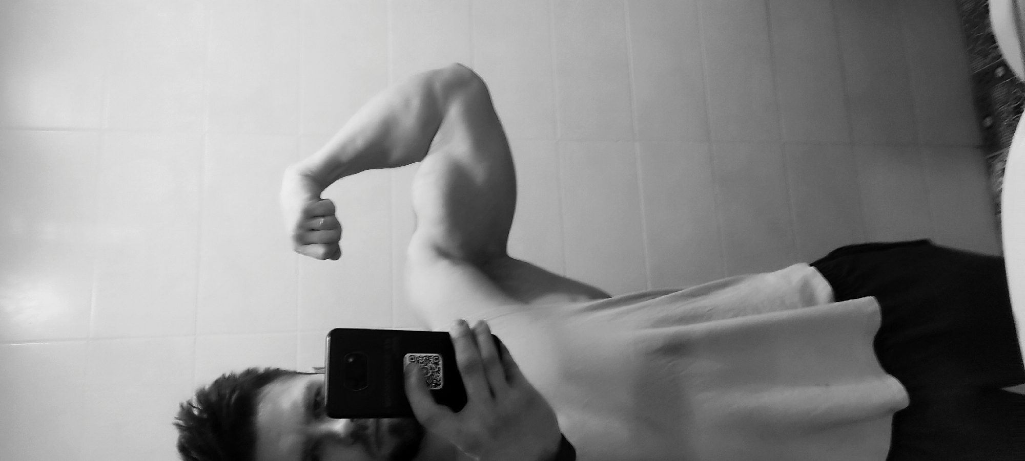 A grayscale image shows a man taking a selfie in what appears to be a bathroom. He is flexing his right bicep, which is prominently displayed in the frame. He is holding a smartphone in his left hand, partially obscuring his face. He is wearing a light-colored t-shirt and dark shorts or pants. The background consists of white square tiles, suggesting a bathroom setting. The location in the city of Fryazino, Russia is not discernible from the image.