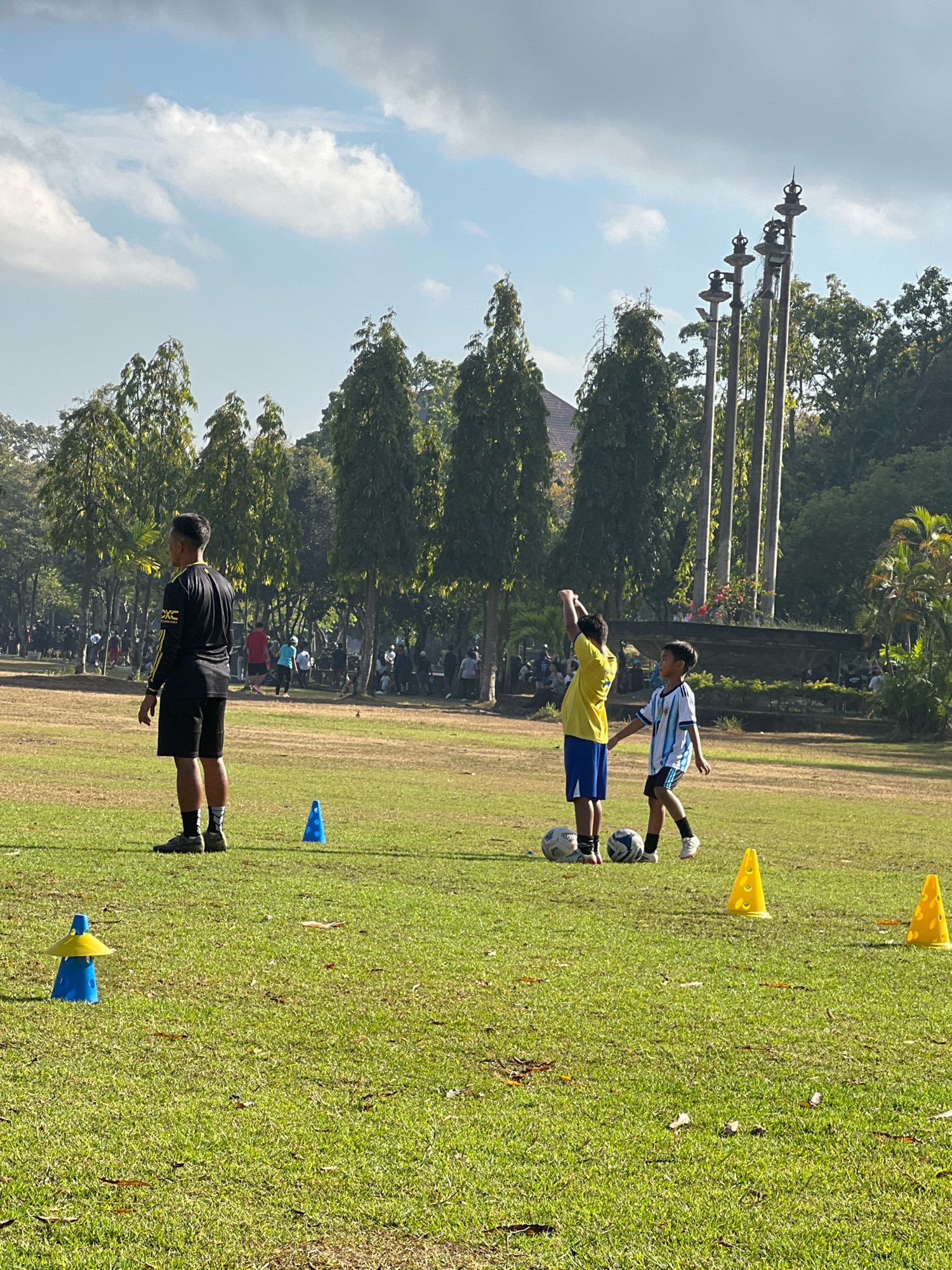 The image captures a sunny day in Denpasar, Indonesia, likely at a park or recreational area, evidenced by the lush green grass, scattered trees, and distant foliage. In the foreground, a man in athletic wear, a black long-sleeved shirt with yellow accents and black shorts, stands facing away from the camera. He is positioned on the grass field, with a bright blue cone to his left.
Further into the field, two young boys are engaged in what appears to be a soccer training session. One boy, wearing a yellow t-shirt and blue shorts, is reaching his arms up. Beside him, a boy in a blue and white striped soccer jersey and black shorts is looking towards the first boy. Two soccer balls are on the ground near them.
In the mid-ground and background, more people are visible, some appearing to be watching or participating in activities. The environment is characterized by a clear blue sky with scattered white clouds. Several tall, slender poles with decorative tops, resembling stylized torches or finials, stand prominently in the background, suggesting a decorative or memorial feature of the location. The overall scene depicts a casual outdoor activity in a well-maintained public space.