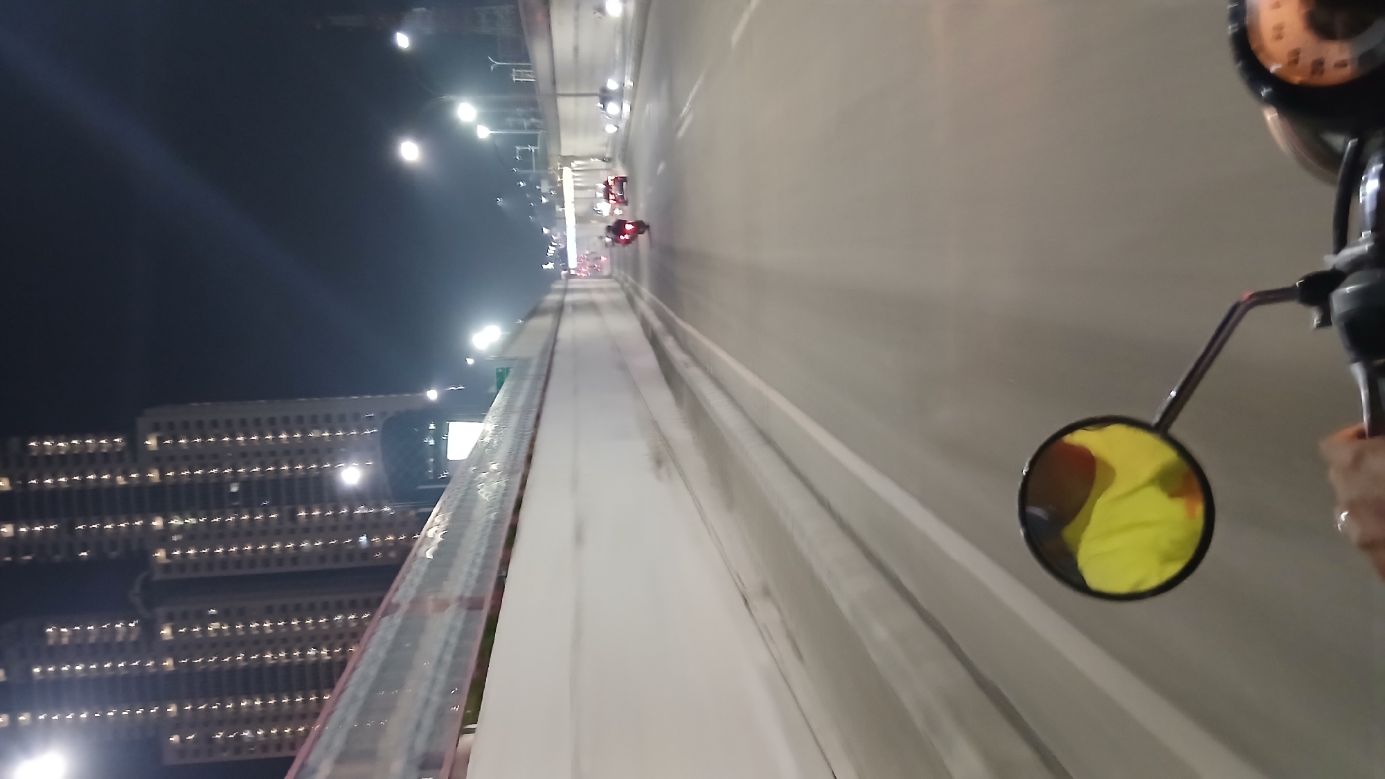A first-person perspective shot shows a motorcycle rider's hand on the handlebars at night, looking forward down a road lined with a tall, white sound barrier wall. In the foreground, a motorcycle's side mirror reflects a person wearing a yellow garment. In the distance, there are several vehicles on the road, including motorcycles and cars, with their headlights and taillights visible. Overhead, streetlights illuminate the scene. In the background, two tall, illuminated apartment buildings stand against the night sky. The scene is set in the City of Medan, Indonesia.