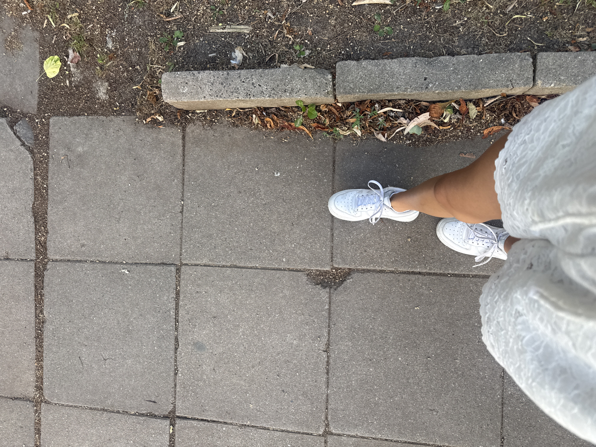 A first-person perspective shot shows a person walking on a paved sidewalk with square gray concrete slabs. The person is wearing white sneakers and a white lacy dress. To the right of the sidewalk, there is a dirt area with some dry leaves and small green plants, bordered by two gray concrete curbs. The lighting suggests it is daytime. The scene is likely in a city like Sofia, Bulgaria, given the architecture of the sidewalk and curbs.