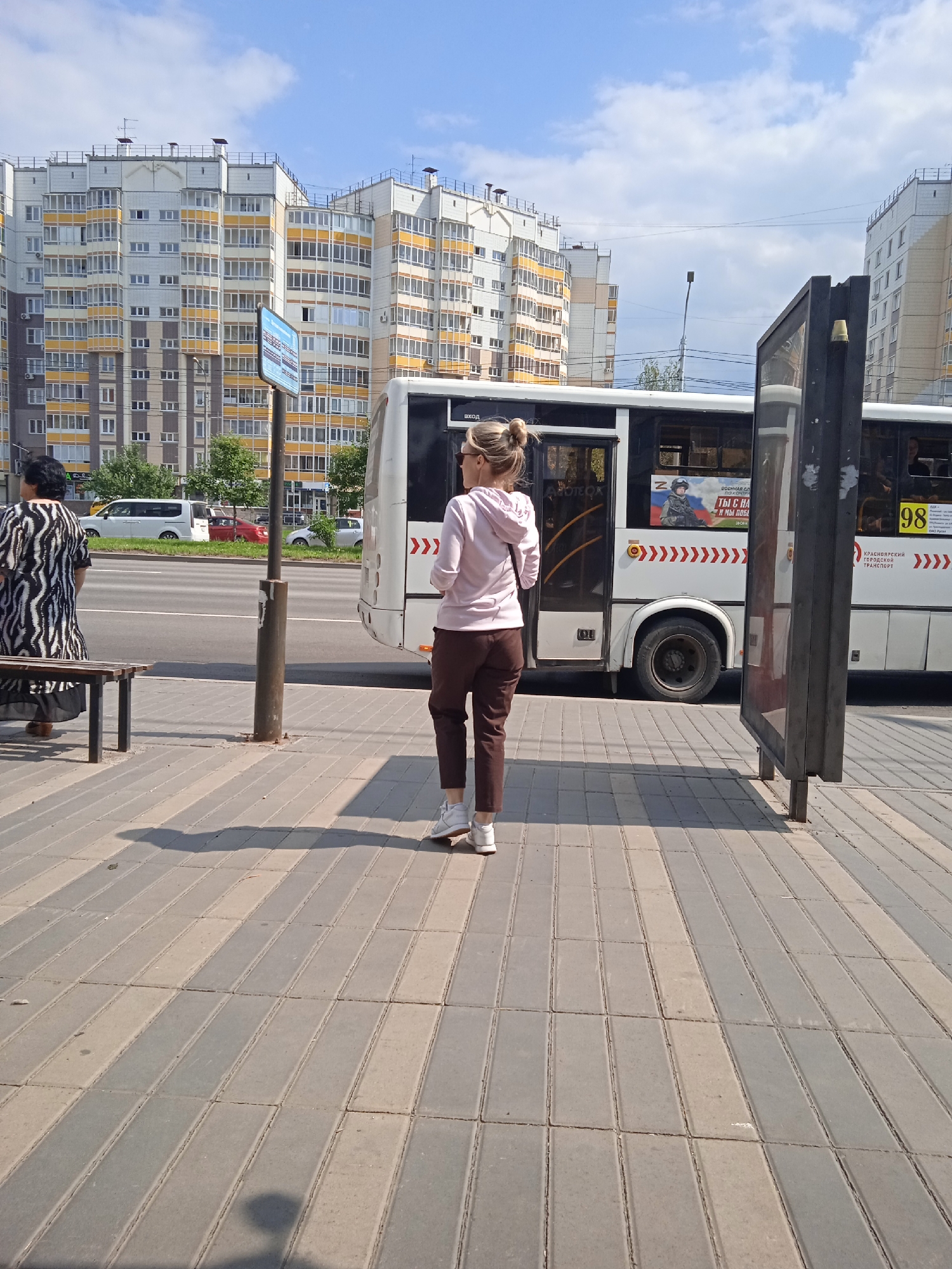 A woman with blonde hair in a bun, wearing a pink hoodie and brown pants, stands on a sidewalk facing a bus. Another woman in a black and white patterned dress is seated on a bench to her left. A white bus with advertisements on its side is parked at the curb. Behind the bus are tall, modern apartment buildings with many windows. The sidewalk is paved with grey and beige rectangular stones. A bus stop sign is visible behind the seated woman. The scene is outdoors on a sunny day. This location is likely Krasnoyarsk, Russia, as indicated by the text on the bus which reads "КРАСНОЯРСКИЙ ГОРОДСКОЙ ТРАНСПОРТ" (Krasnoyarsk City Transport).