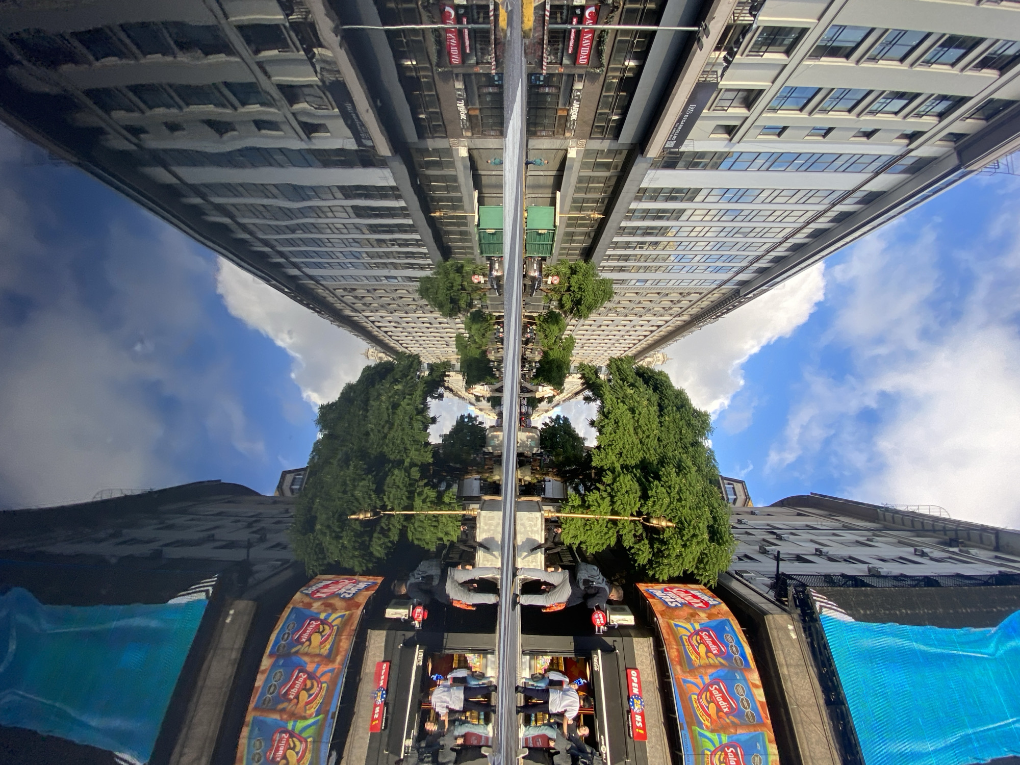 The image displays an urban street scene, likely in Buenos Aires, Argentina, characterized by a striking horizontal reflection that creates a unique symmetrical composition.
A dark, highly reflective surface runs horizontally across the middle of the frame, effectively dividing the image. The **upper half** presents an inverted reflection of tall city buildings and a bright blue sky with scattered white cumulus clouds. Architectural details and numerous windows are visible in this mirrored segment.
The **lower half** shows the direct view of the street-level environment. Here, tall, modern buildings with many windows rise from the bottom edge towards the upper right. Lush green trees, with dense foliage, are interspersed along what appears to be a median or sidewalk. At the very bottom of the frame, several people are visible walking or standing, suggesting typical urban pedestrian activity. Large, colorful billboards or digital screens are prominently displayed, showcasing advertisements.
The scene takes place during the daytime under a clear sky with scattered clouds. The intense reflection is a notable detail, making the street or a large architectural surface act like a mirror. The composition highlights the verticality of the buildings and the dynamic symmetry created by the reflection.
Visible text includes: "AS DESARROLLOS" on a building, "NAVIDAD CAS" (appearing inverted in the reflection), and "JAROK" or "JAROK LOK" on signs. Billboards prominently feature "Saladix," a snack brand, alongside a sign reading "OPEN 25 HS!". Distorted reflections of "Saladix" such as "xibala?" and "Zajaqix" are also discernible.