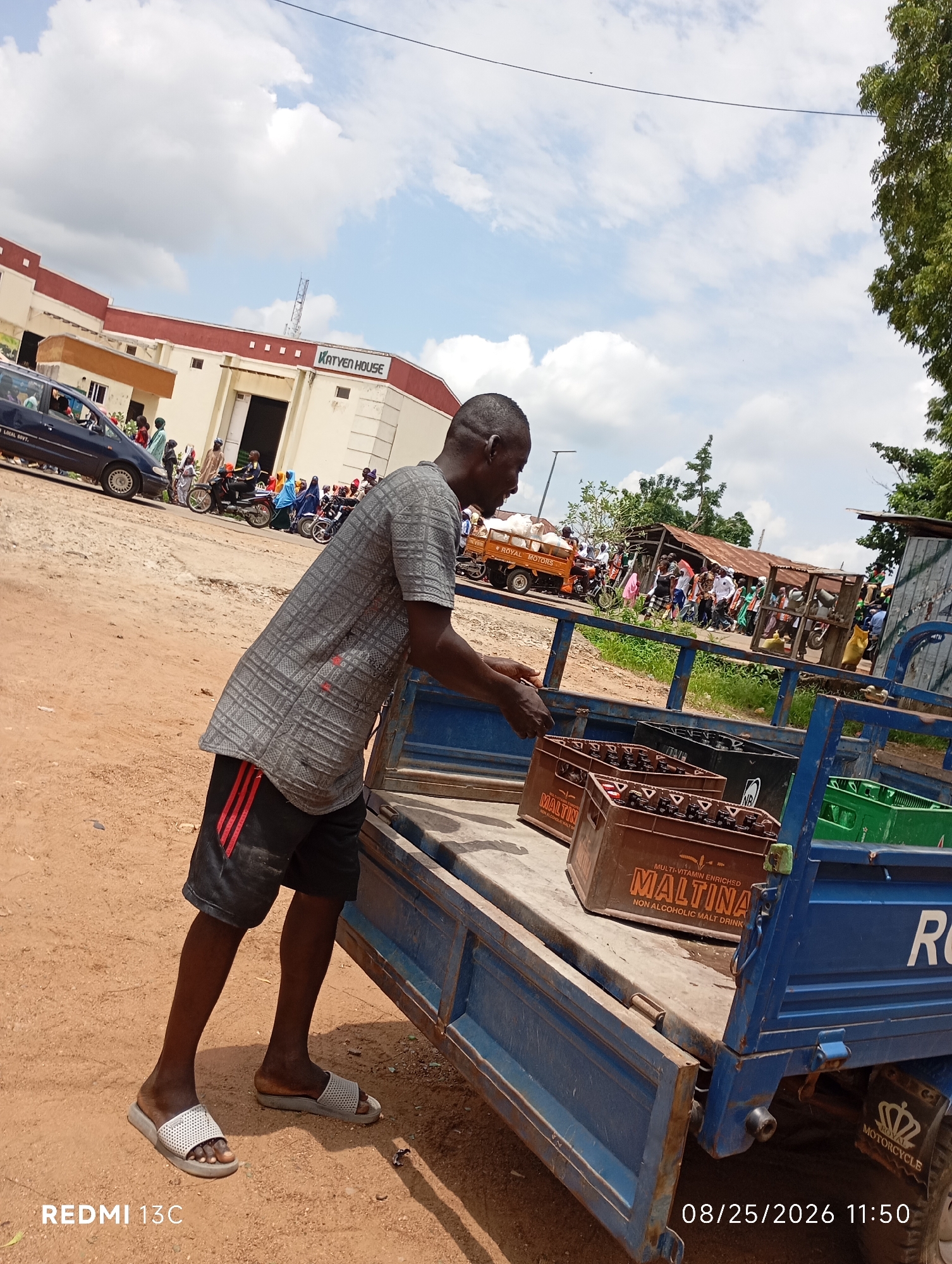 This image captures a scene in Shendam, Nigeria, on August 25, 2026, at 11:50 AM. The central figure is a dark-skinned man in a grey patterned short-sleeved shirt and black shorts with red stripes, wearing grey slides. He is standing on a dusty, unpaved road, next to the open back of a blue, three-wheeled utility vehicle. The vehicle's bed is partially filled with brown crates of "Maltina" non-alcoholic malt drink and green plastic crates.
In the background, there is a light-colored building with a maroon trim and the inscription "KATVEN HOUSE." Several people are gathered around, some on motorcycles and others on foot, suggesting a public gathering or marketplace. A dark blue car is parked to the left, and further in the background, there are some makeshift structures with corrugated iron roofs, trees, and vegetation. The sky is partly cloudy. The overall impression is a bustling, everyday scene in a Nigerian town.