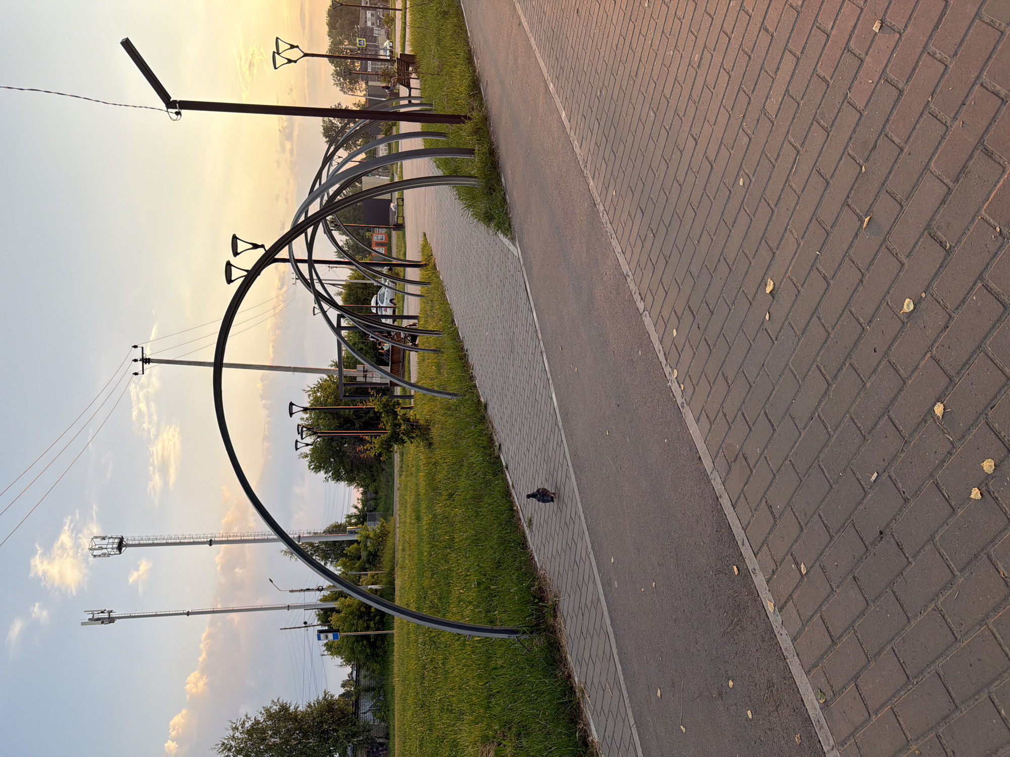 An outdoor scene depicts a pathway designed with a series of connected metal arches, creating a tunnel-like effect. The pathway itself is divided into two distinct surfaces: one paved with brick-like stones and the other a smoother, darker asphalt. Along the side of the pathway, a stretch of green grass is visible, bordered by trees. In the background, two tall cellular towers stand prominently against a sky with scattered clouds, illuminated by the warm glow of either sunset or sunrise. Several streetlights, designed with a modern, minimalist aesthetic, are positioned along the pathway. A pigeon is seen walking on the brick-paved section of the pathway. The overall environment suggests a public park or a recreational area within the city of Chaikovsky, Russia, given the architectural style and the presence of communication towers typical of urban infrastructure.