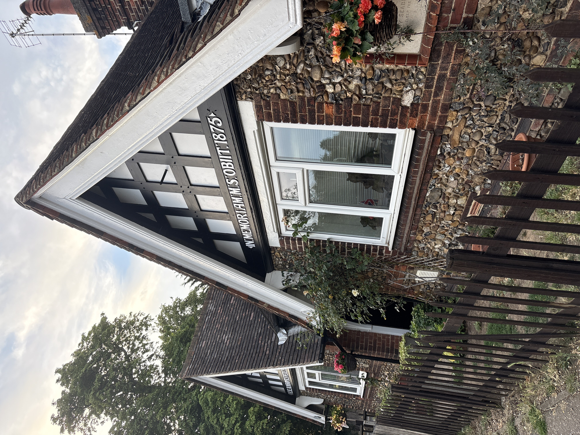The image shows a section of two attached Tudor-style cottages in Chelmsford, United Kingdom. The cottages are constructed with red brick and stone foundations, featuring dark timber framing on their gabled roofs. Each gable displays a white fascia board with dark timber infills. The left cottage has a small window with a hanging flower basket to its left. The right cottage has a larger window and a hanging basket with orange flowers to its right. Above the right cottage's window, a black sign with white lettering reads "IN MEMORIAM M. S. OBIIT. 1875". A dark wooden picket fence runs along the front of the properties, separating them from the street. Trees and a cloudy sky are visible in the background. There are no people or animals visible in the scene.