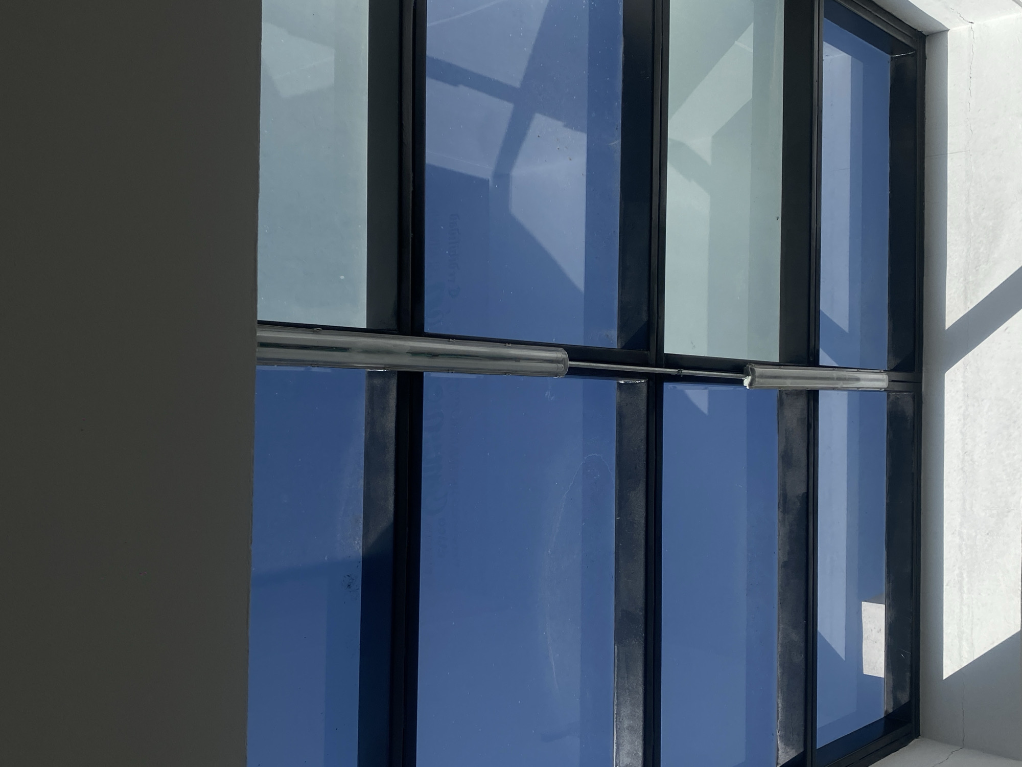A close-up, vertical-oriented architectural photograph captures a section of a building featuring large windows with dark frames. The windows primarily reflect a bright blue sky, suggesting a clear, sunny day. Some panes also reflect lighter, possibly cloud-filled or structural elements, creating abstract geometric shapes within the glass.
A prominent, horizontal, silver-colored metallic bar or pipe extends across the middle of two window panes, positioned approximately at the center height of the image.
On the left side, a flat, light grey wall or column partially frames the window section. On the right, a textured, off-white wall is visible, showing signs of wear or cracking, and casting sharp, angular shadows from an unseen structure to the right, further indicating strong sunlight.
Notable details include a faint, reversed reflection of text on one of the blue window panes, appearing to spell "Coca-Cola Light" or "Coca-Cola Lift". Additionally, a small, rectangular white sticker or label is affixed to the lower right window pane. The overall scene is static, capturing the interplay of light, reflection, and architectural forms.