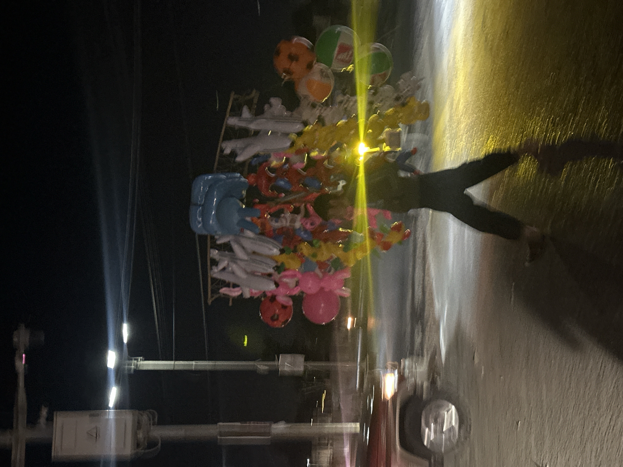 At night in Quetta City Tehsil, Pakistan, a street vendor carries a large assortment of balloons, toys, and figurines, while walking alongside a roadway. The collection includes character balloons (likely Spiderman), small airplane figures, and several round balloons. A shadow of the vendor is cast on the bright pavement, indicating the presence of streetlights or vehicle headlights. Traffic signals, utility poles, and a vehicle with headlights are visible in the background, suggesting a moderately busy urban street.