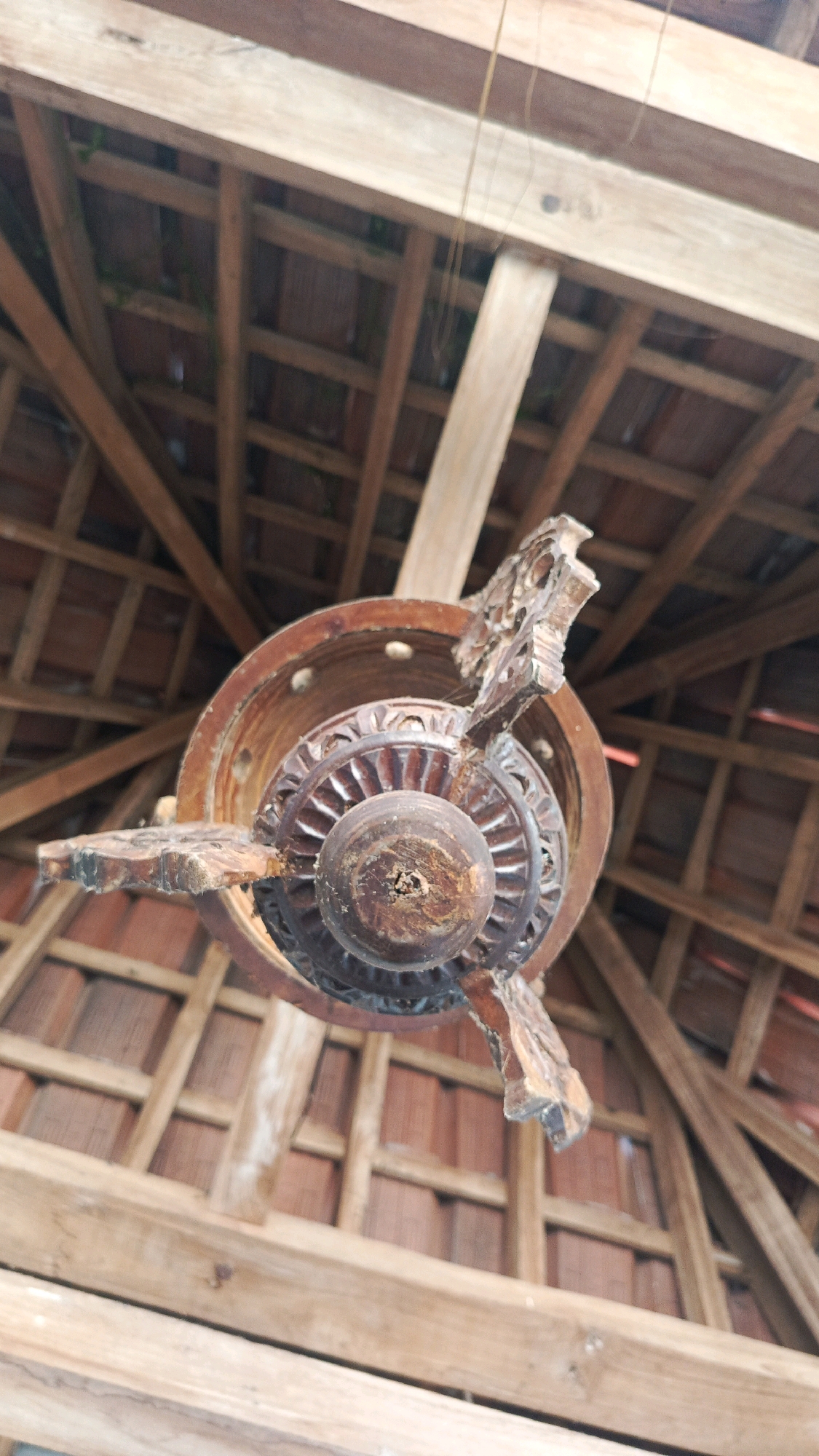 The image shows the underside of a wooden ceiling with exposed beams and terracotta tiles. A carved wooden decorative element, possibly part of a lamp or fan, hangs from the center. The scene appears to be outdoors or in a structure with open sides, typical of traditional Indonesian architecture. The location is identified as Babadan, Indonesia, which is a village in the Madiun Regency of East Java. The image is captured from a low angle, looking upwards towards the ceiling structure. There are no people or animals visible in the image, and no actions or interactions are taking place.