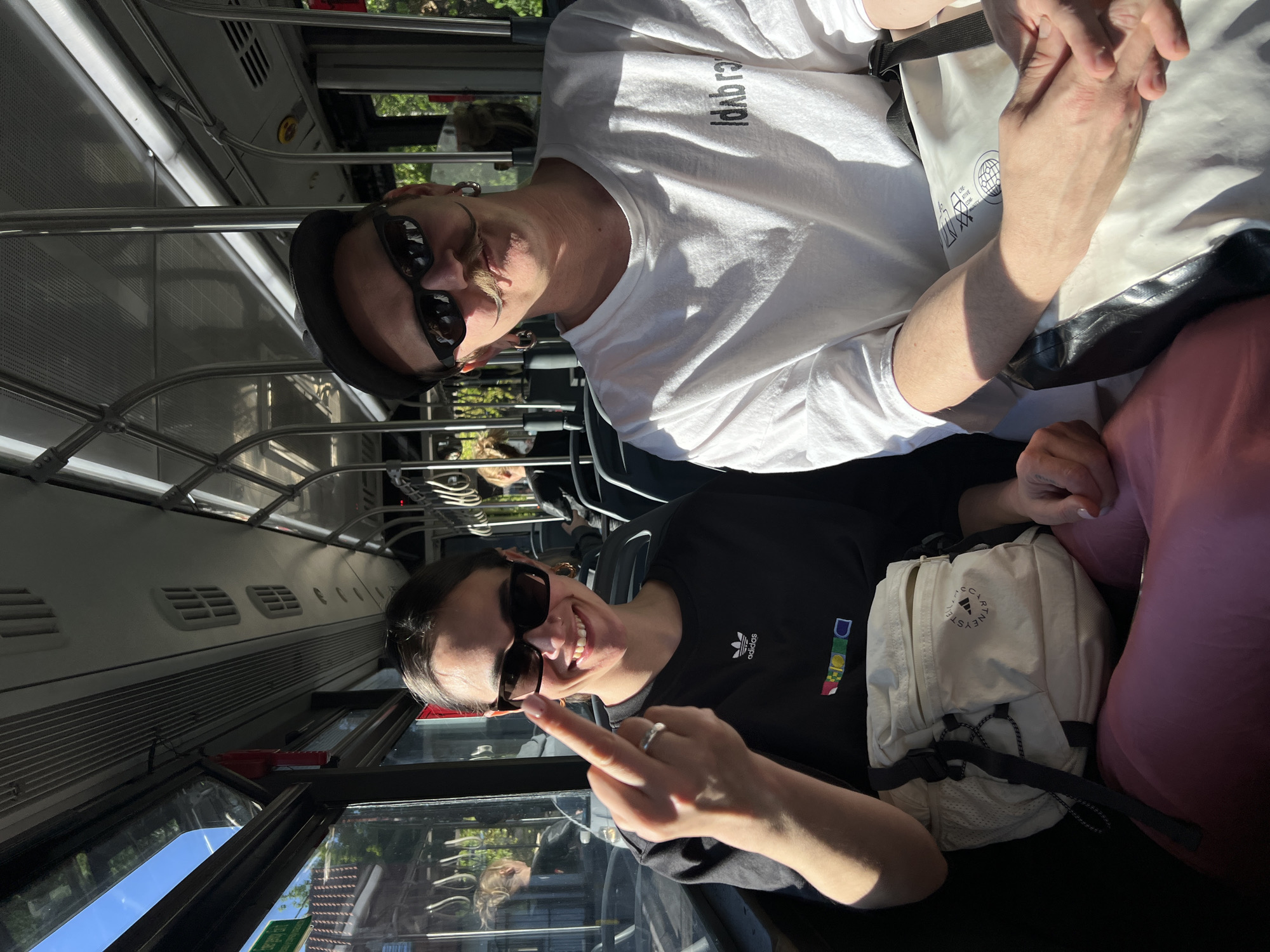 Two individuals are seated on a moving vehicle, likely a bus, based on the interior. The woman in the foreground, wearing a black Adidas t-shirt and sunglasses, is giving the middle finger to the camera. A white backpack is situated near her. The second individual in the background is wearing a white t-shirt with black text and sunglasses. The bus interior features metal railing and rows of seats. Through the window, greenery and buildings are visible, suggesting an urban environment. This photo was most likely taken in Vozdovac Urban Municipality, Serbia.