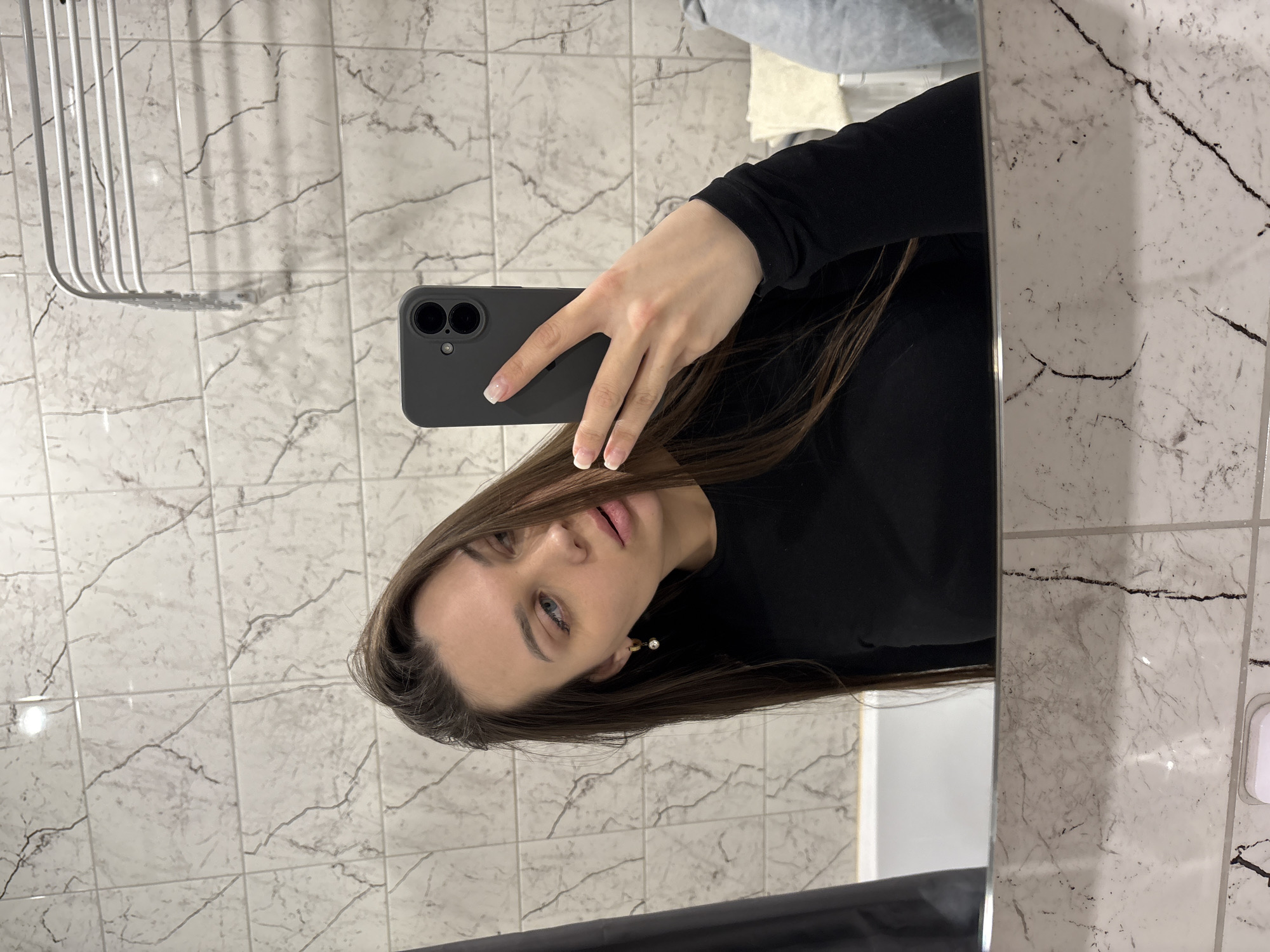 An indoor mirror selfie captures a woman holding a dark smartphone to take her picture. The image is rotated 90 degrees clockwise from a typical upright view.
**Subjects:**
The primary subject is a young woman with long, straight, dark brown hair, parted slightly to her right (viewer's left). She is wearing a black long-sleeved top. Her visible hand, holding the phone, has a light-colored French manicure. Her face is turned slightly towards the camera, revealing light-colored eyes (possibly blue or grey) with visible mascara, and her lips are slightly parted. A delicate, light-colored earring is visible on her right earlobe. She is looking directly into the phone's lens, which is reflected in the mirror.
**Objects:**
She is holding a dark gray or black smartphone with two prominent circular camera lenses on its rear, encased in a simple dark cover.
In the background, to the left of her reflection, a metallic, multi-barred rack or shelf is partially visible.
**Scene and Setting:**
The setting appears to be an indoor room, likely a bathroom, characterized by the light-colored square tiles on the wall. These tiles have a marbled pattern with dark, irregular veins running through them. The reflections in the mirror suggest bright, diffused indoor lighting.
**Activity:**
The woman is actively taking a mirror selfie portrait.
**Notable details:**
Her expression is neutral, almost contemplative. The tiling suggests a clean, modern interior design. There is no visible text in the image. The time of day is not discernible, as the scene is illuminated by artificial indoor lighting. Given the context, the location is Yekaterinburg, Russia.