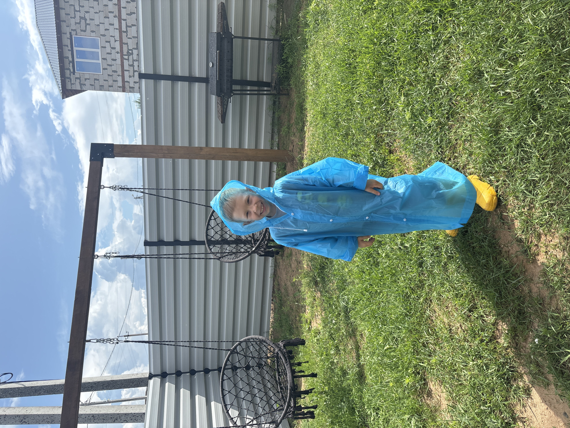 A young boy, wearing a bright blue hooded raincoat and yellow boots, stands on a grassy lawn. He is smiling at the camera. In the background, there is a grey metal fence, two black woven hanging chairs, and a wooden swing set frame. To the right of the swing set, a black barbecue grill is visible. A partially constructed brick building with a pitched roof and a window is situated behind the fence. The sky above is blue with scattered white clouds. This scene appears to be in a residential backyard in Chaikovsky, Russia, given the construction style of the building and the typical suburban landscape.